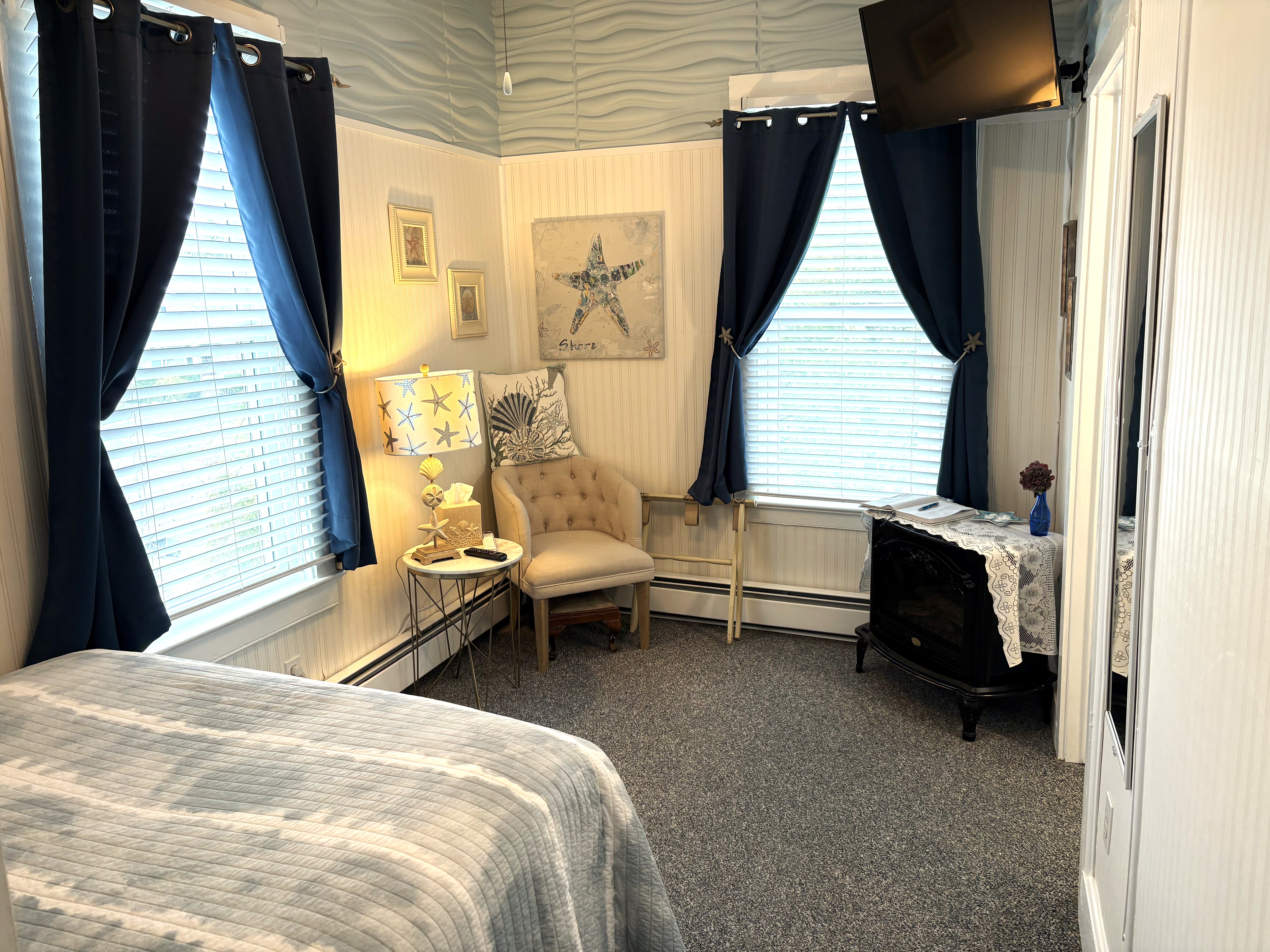 A cozy bedroom with blue curtains, a reading chair, an electric wood stove, a wall-mounted TV, and nautical-themed decor including a starfish lamp and wall art.