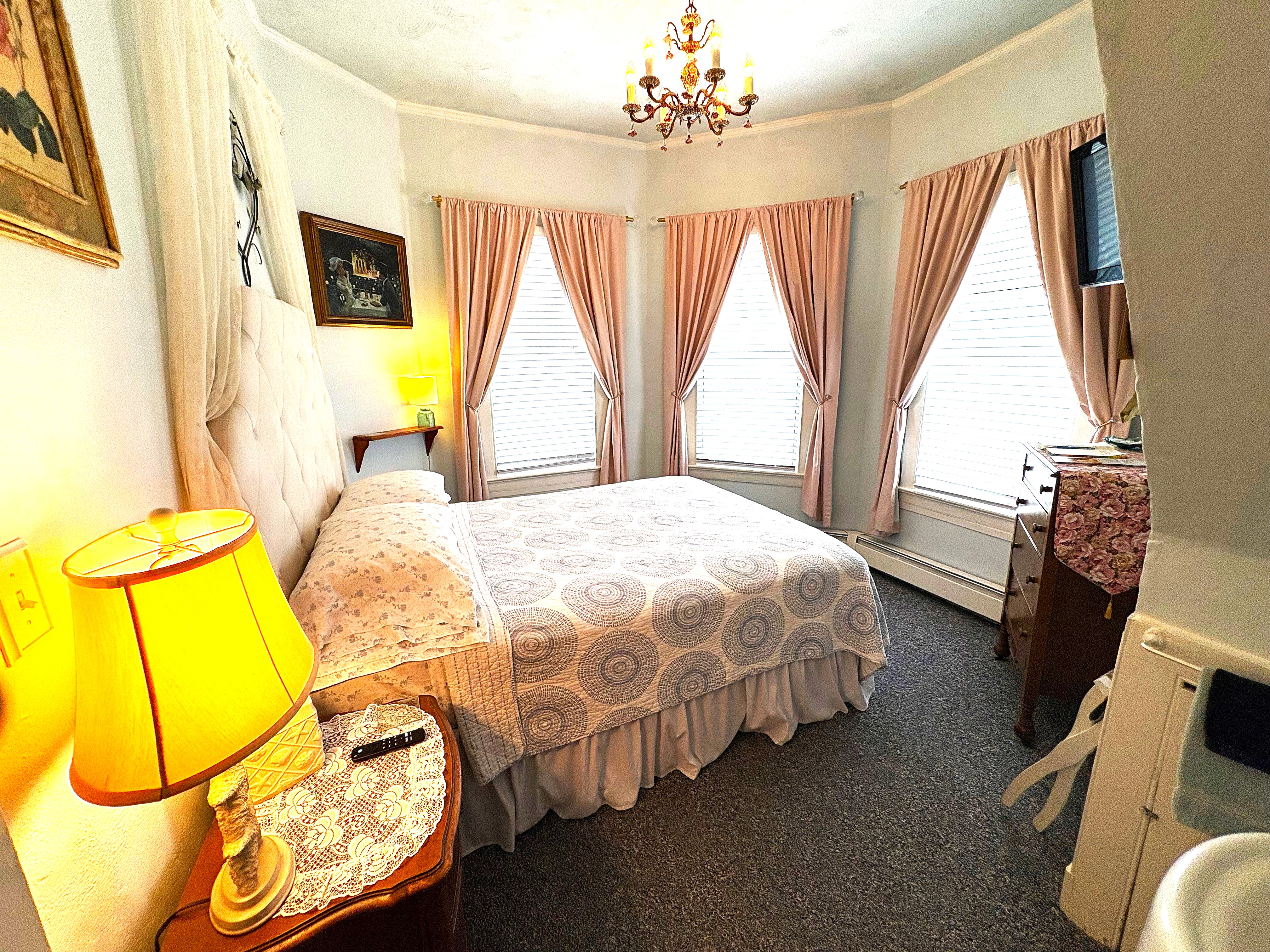 An elegant guest room with a queen bed, a classic crystal chandelier, and large windows with soft pink curtains. A vintage lamp sits on a bedside table in the foreground.