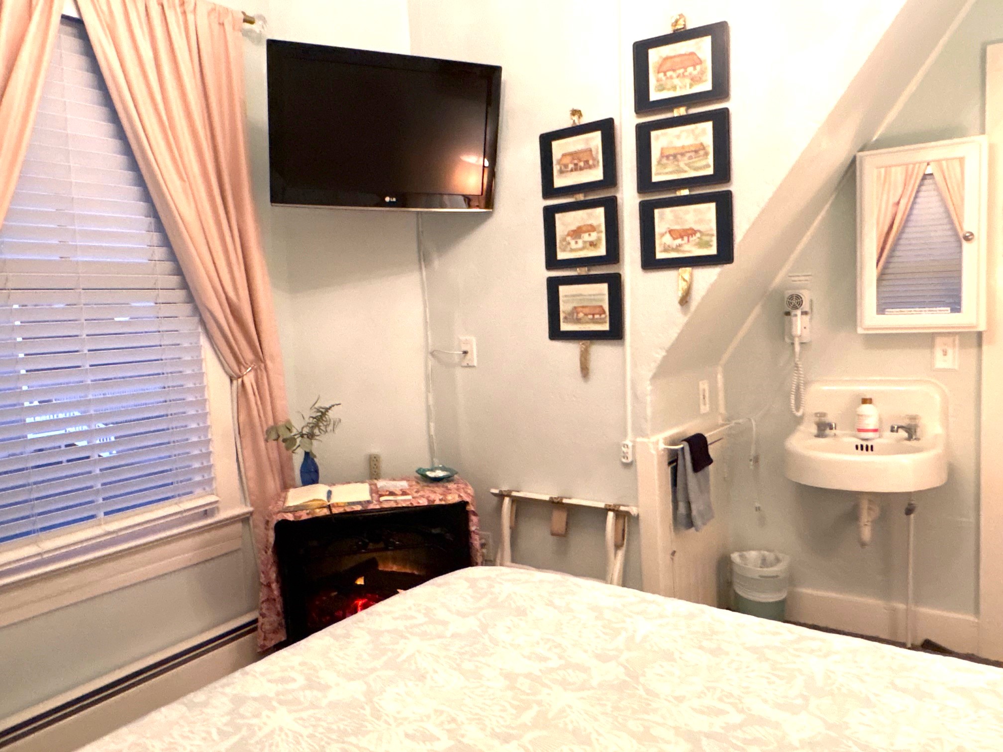 An interior view of a charming guest room featuring a wooden dresser topped with a small vase, a wall-mounted television, and a vintage-style wall-mounted sink with a mirrored cabinet.