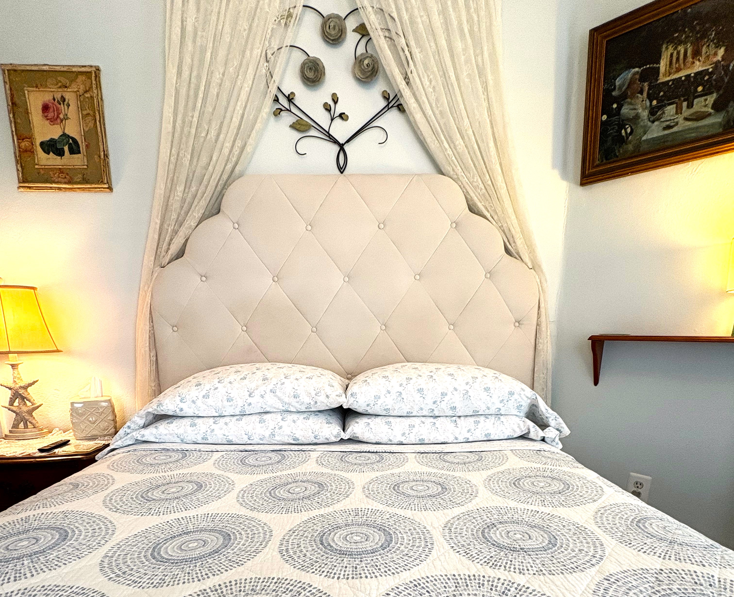 An interior view of a bright guest room featuring a bed with a tufted white headboard and patterned gray bedding. Above the headboard, a white sheer canopy is draped over a decorative wall ornament. To the left, an open door reveals a bathroom with a white shower curtain.