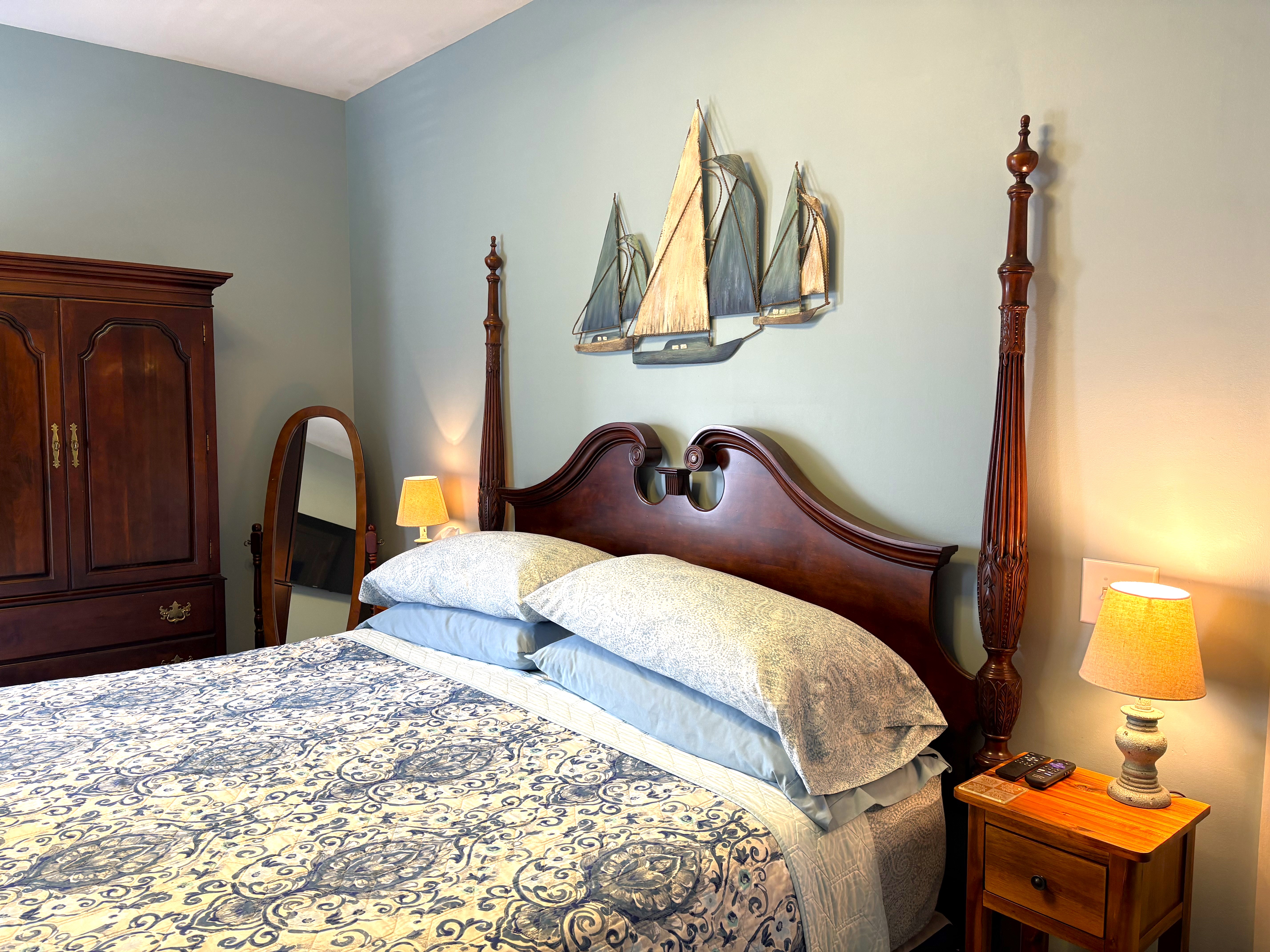 An interior view of a bedroom featuring a dark wood four-poster bed with a curved headboard, a blue and white patterned comforter, and a large metal wall hanging of three sailboats above the bed.