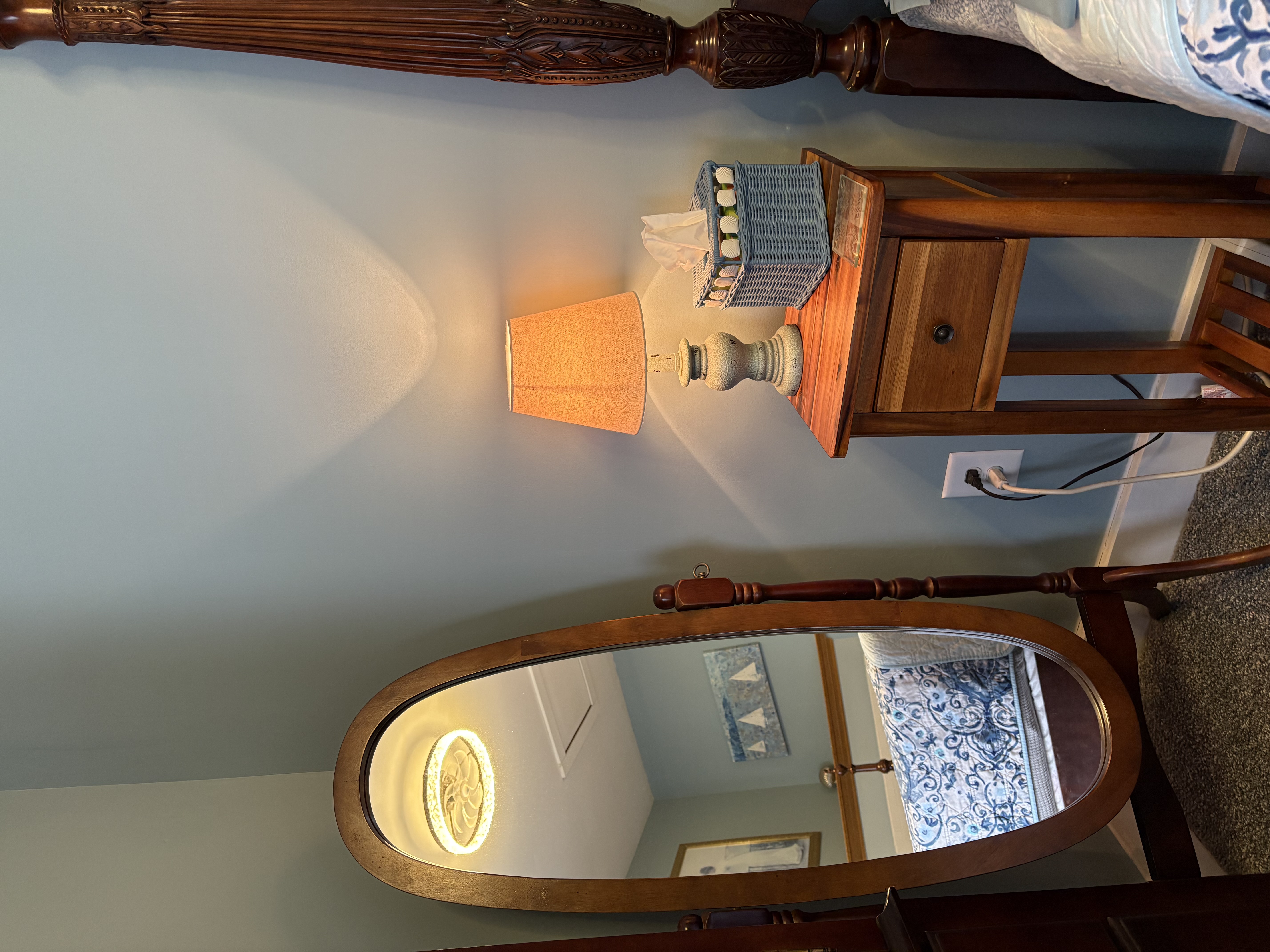 An interior view of a bedroom corner featuring a full-length oval dressing mirror on a wooden stand, an antique-style wooden nightstand with a lit lamp, and the post of a dark wood bed frame.