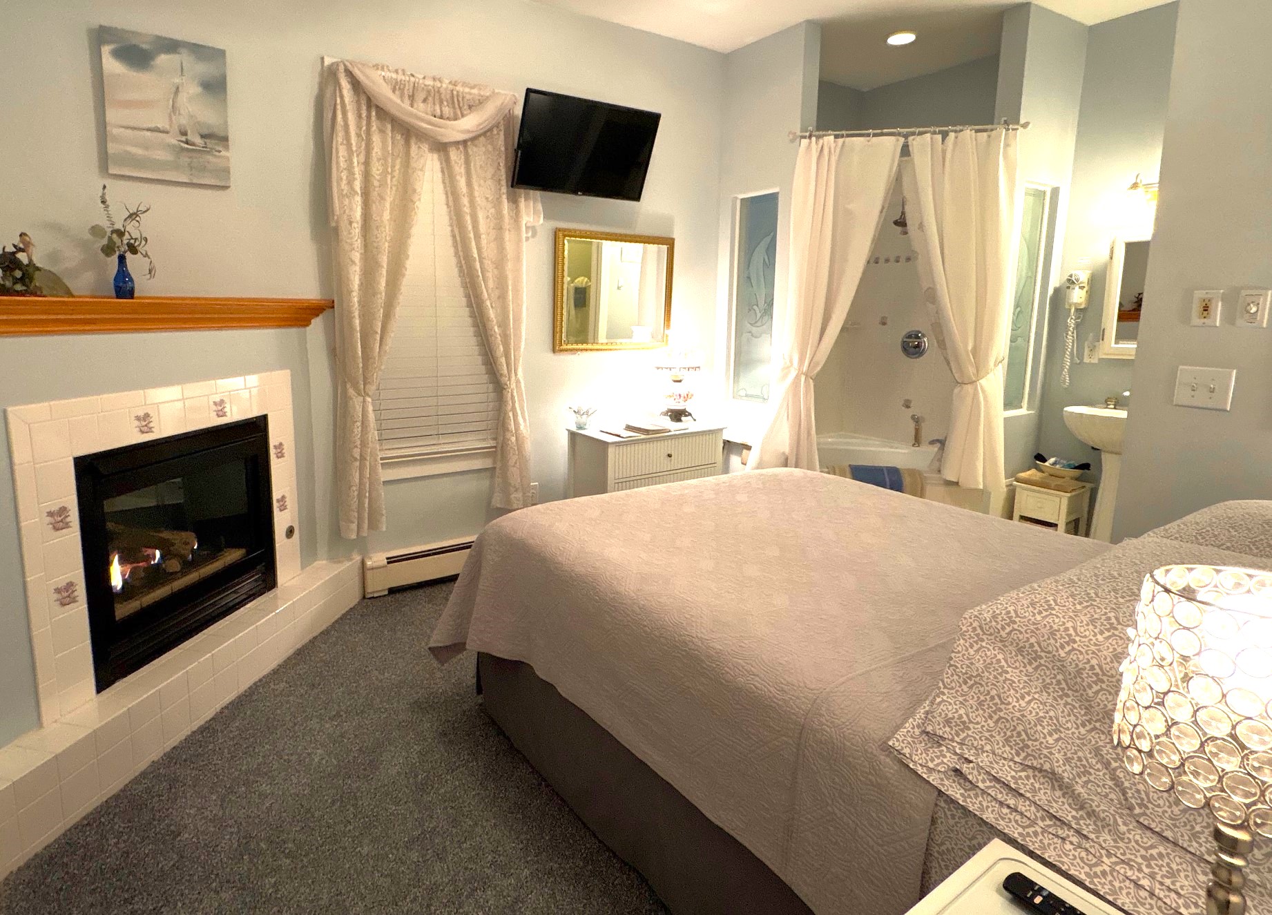 An interior view of a bright guest room featuring a plush gray tufted headboard, a bed with a circular patterned quilt, and a built-in corner fireplace with a white mantel and decorative tiles.