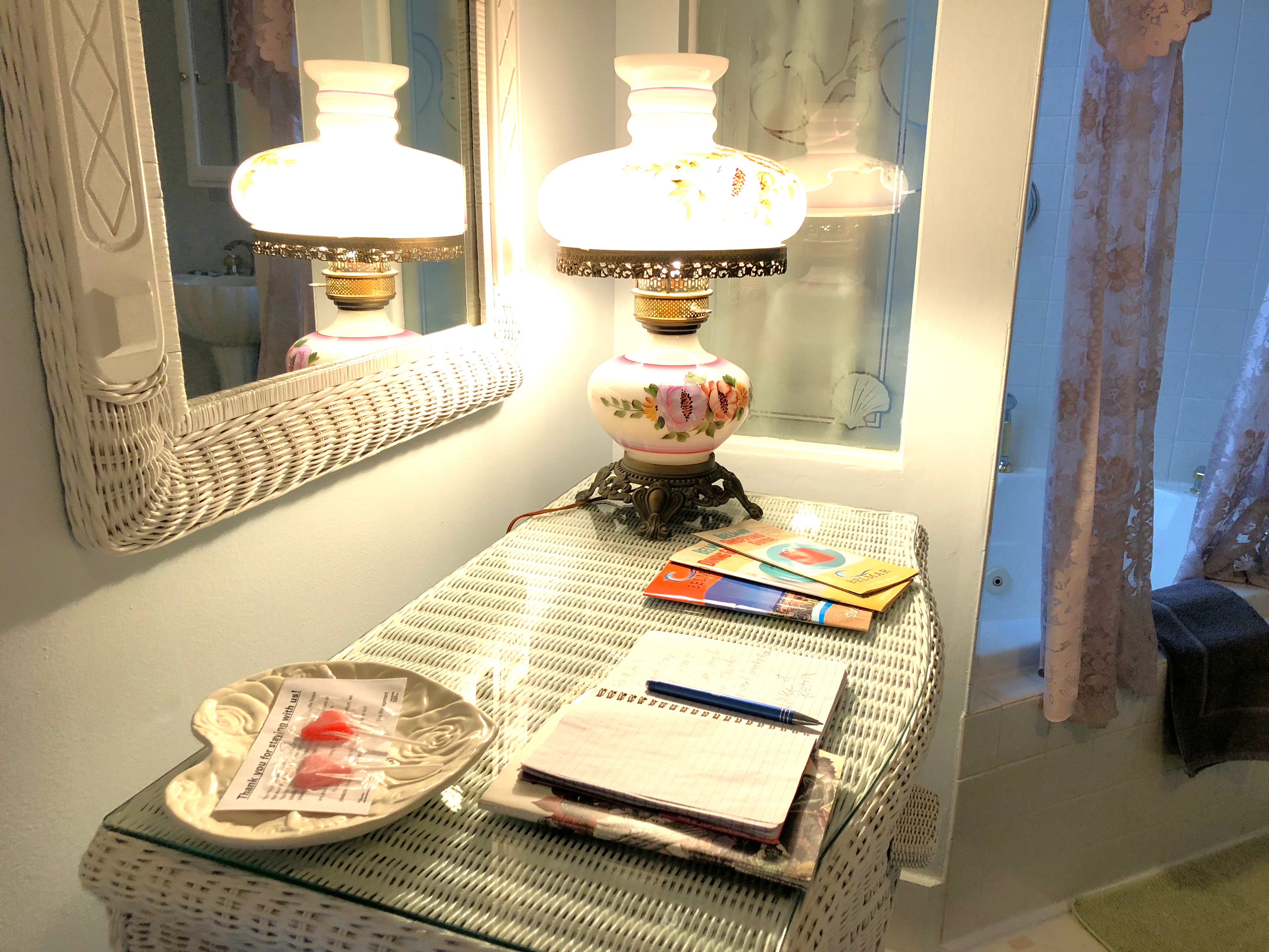 A close-up view through the glass of a lit gas fireplace, showing glowing blue and orange flames flickering around ceramic logs, with the corner of a bed and a guest room reflected in the glass.