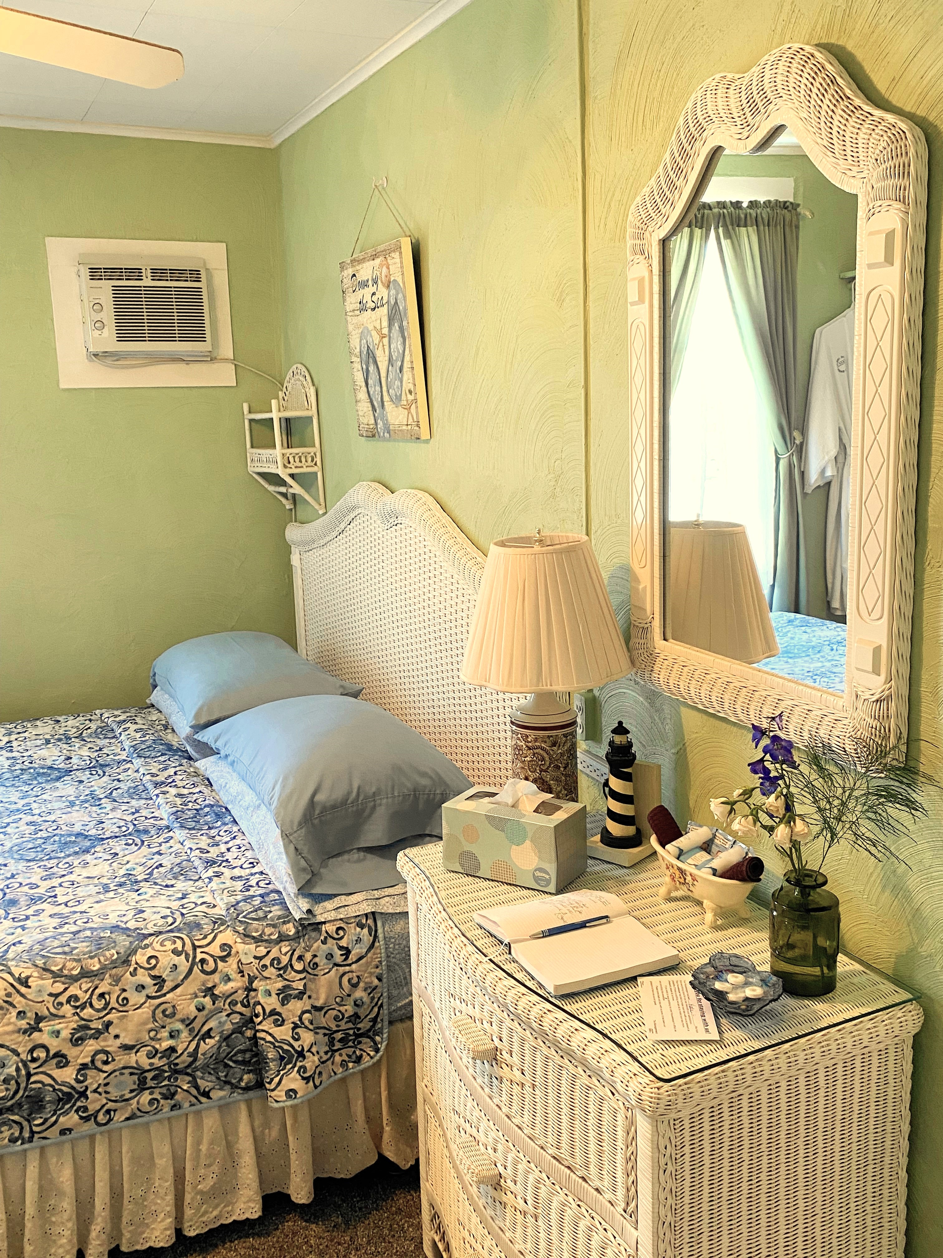 An interior view of a bright guest room with light green walls, featuring a bed with a white headboard and blue patterned bedding. A white wicker dresser in the foreground holds a guest book, a lighthouse-themed lamp, and a vase of flowers, with a matching wicker-framed mirror mounted on the wall above it.