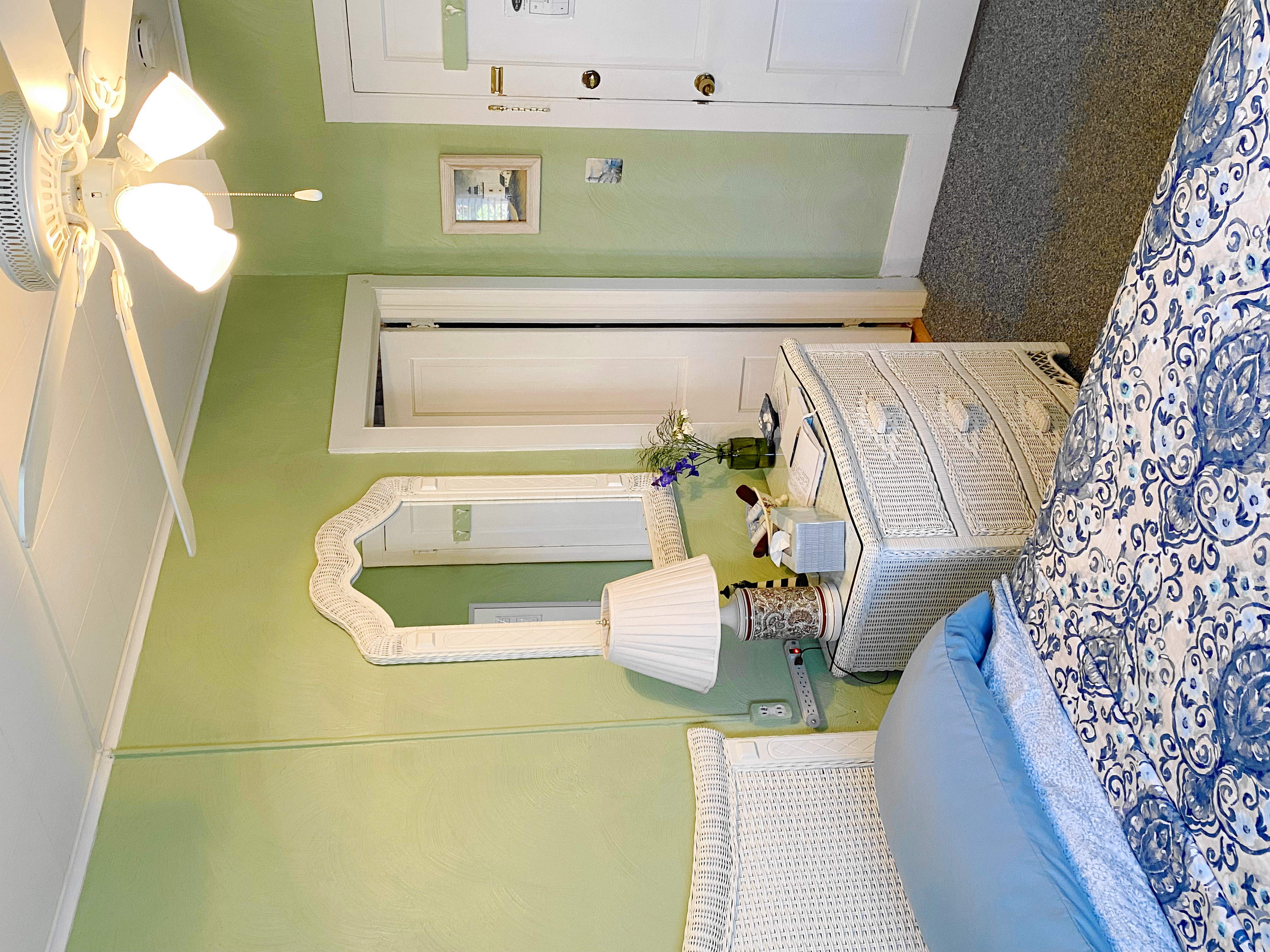 An interior view of a bedroom with light green walls featuring a bed with a white wicker headboard and blue patterned bedding. A small white wicker wall shelf hangs above the bed next to a piece of coastal wall art depicting flip-flops on a sandy beach.