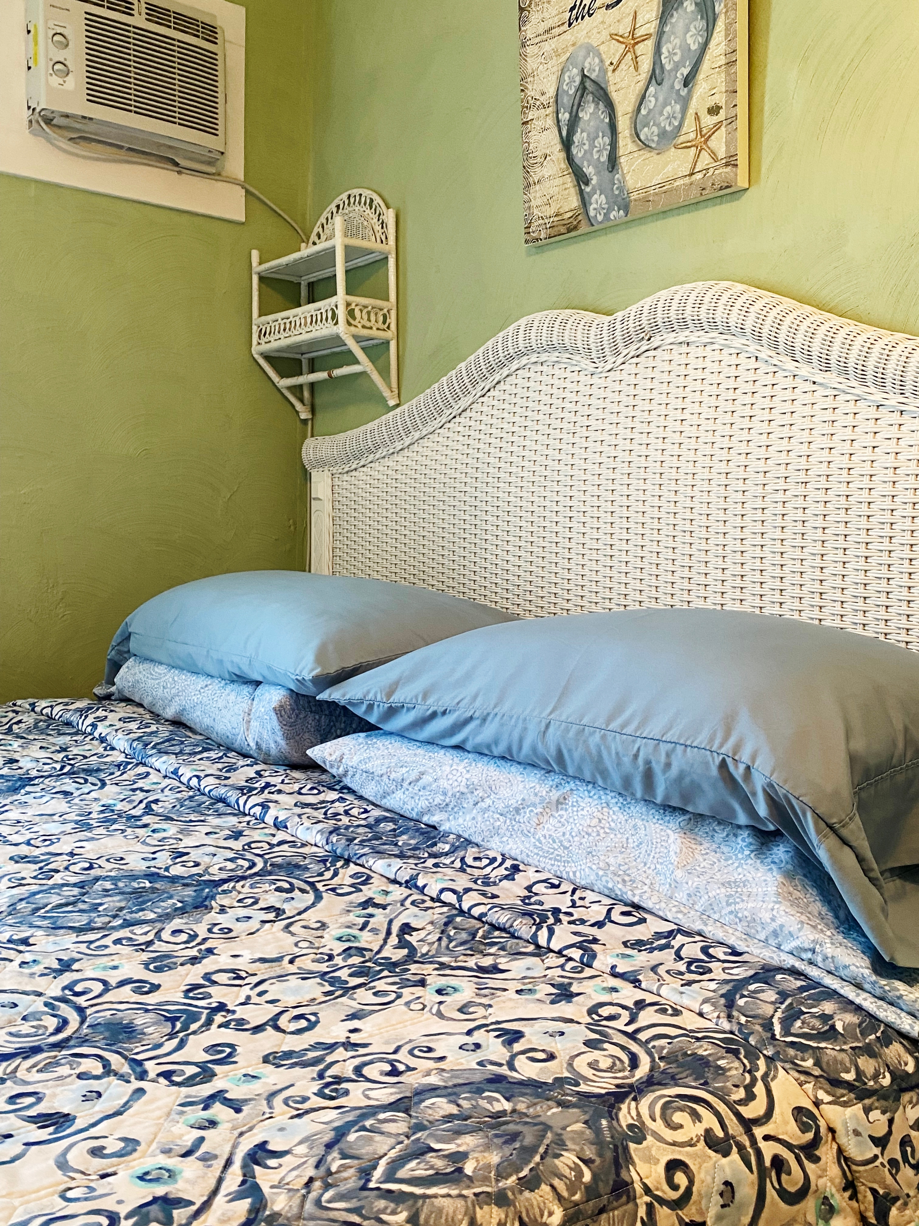 An interior view of a bright guest room with sage green walls, featuring a bed with a blue and white patterned quilt. A white bathrobe hangs from a wall-mounted rack next to a window with olive green curtains, and a lit ceiling fan provides a warm glow from above.