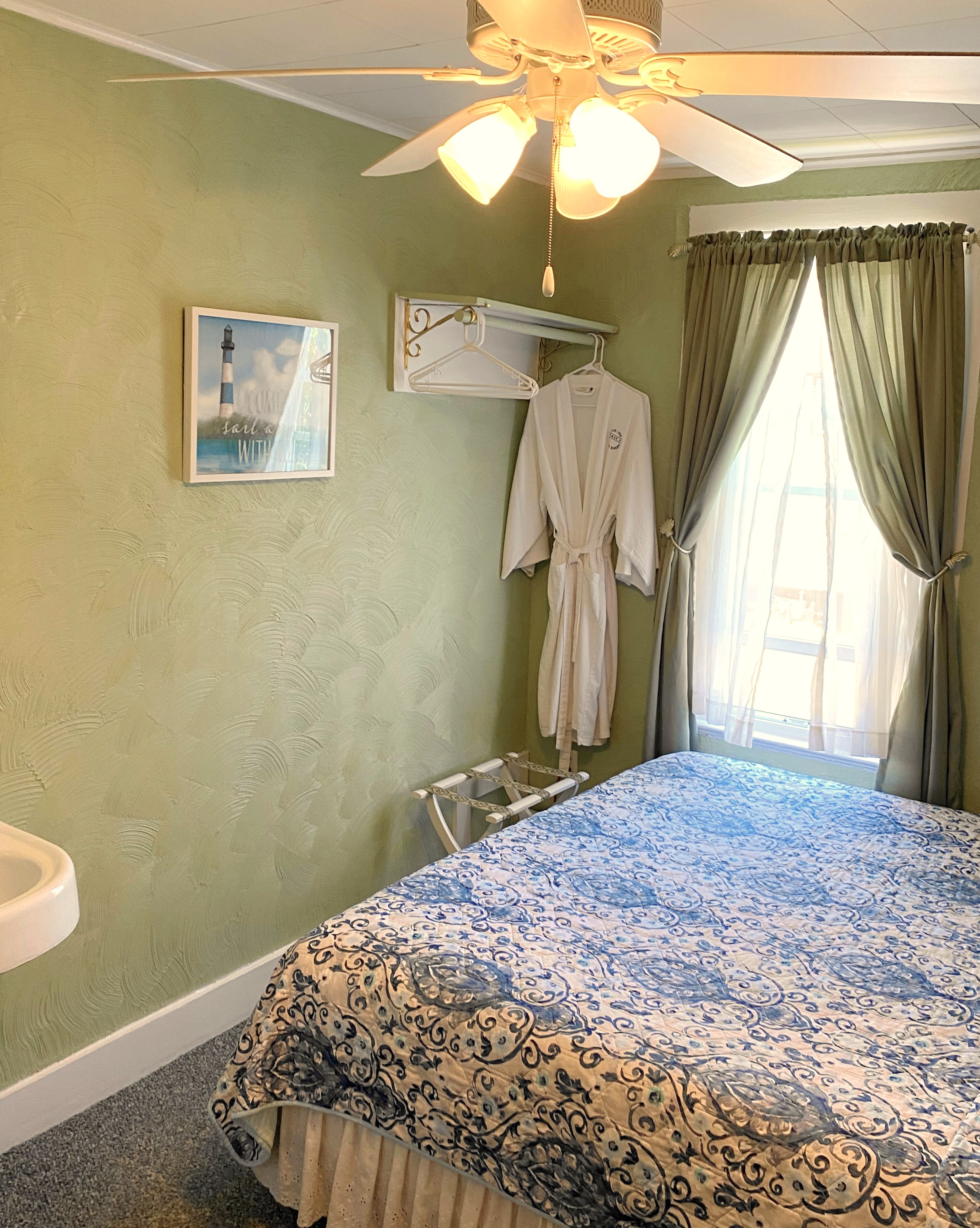 An interior view of a cozy seating area featuring a dark gray sofa with a decorative floral pillow, set against a wall with rustic wood paneling. A small white wicker side table with a modern lamp stands next to the sofa, and an open doorway in the background reveals a glimpse of an adjacent room with light green walls.