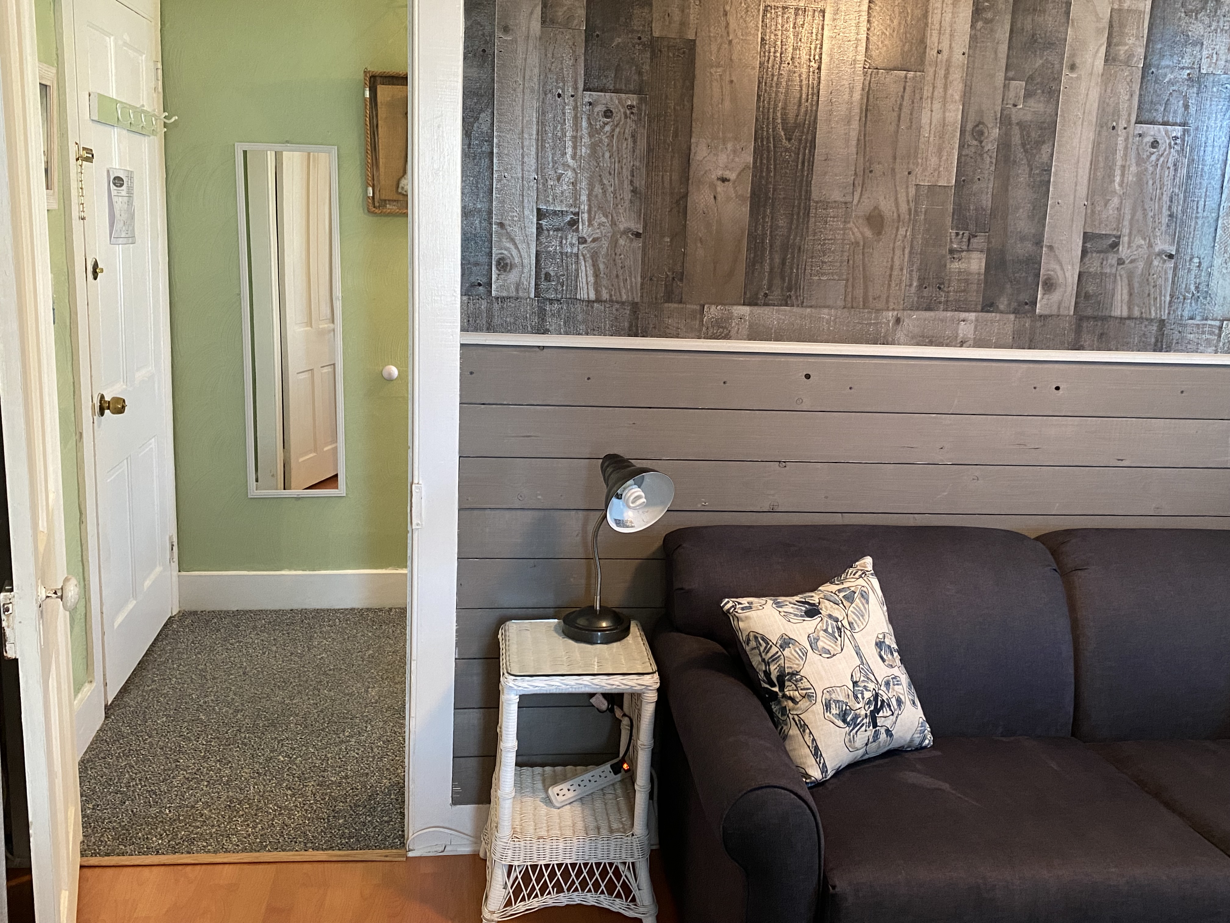 An interior view of a cozy seating area featuring a dark gray sofa with decorative floral pillows, a traditional blue and gray patterned rug, and a small bistro-style table with two stools. A wall-mounted television is positioned across from the sofa, and a lit ceiling fan provides a warm glow over the wood-plank flooring.