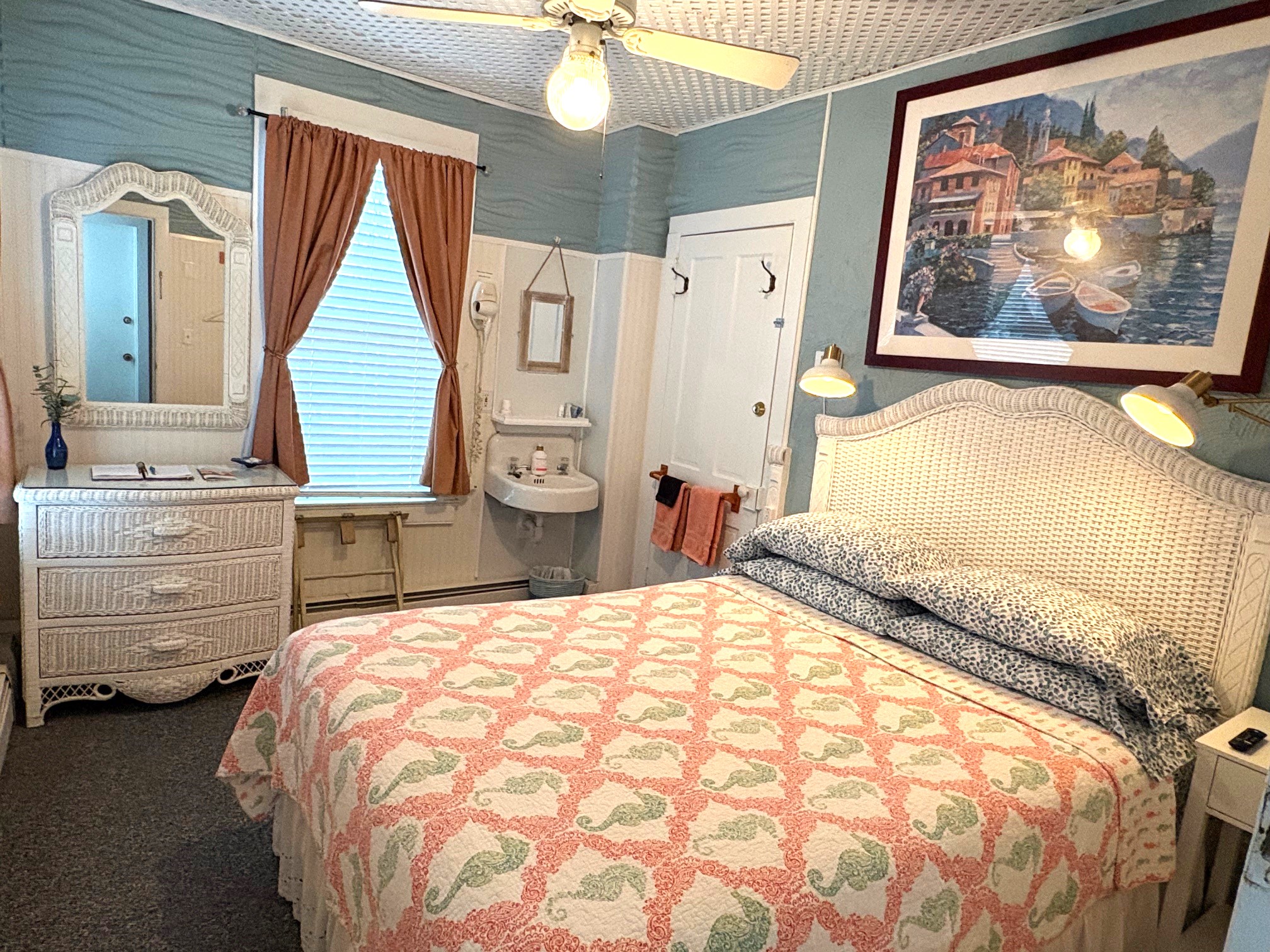 An interior view of a bright guest room with light blue and white walls, featuring a bed with a white headboard and a coral-colored quilt. The room includes a wooden dresser, a small wall-mounted sink, a window with grey curtains, and various framed coastal artworks.