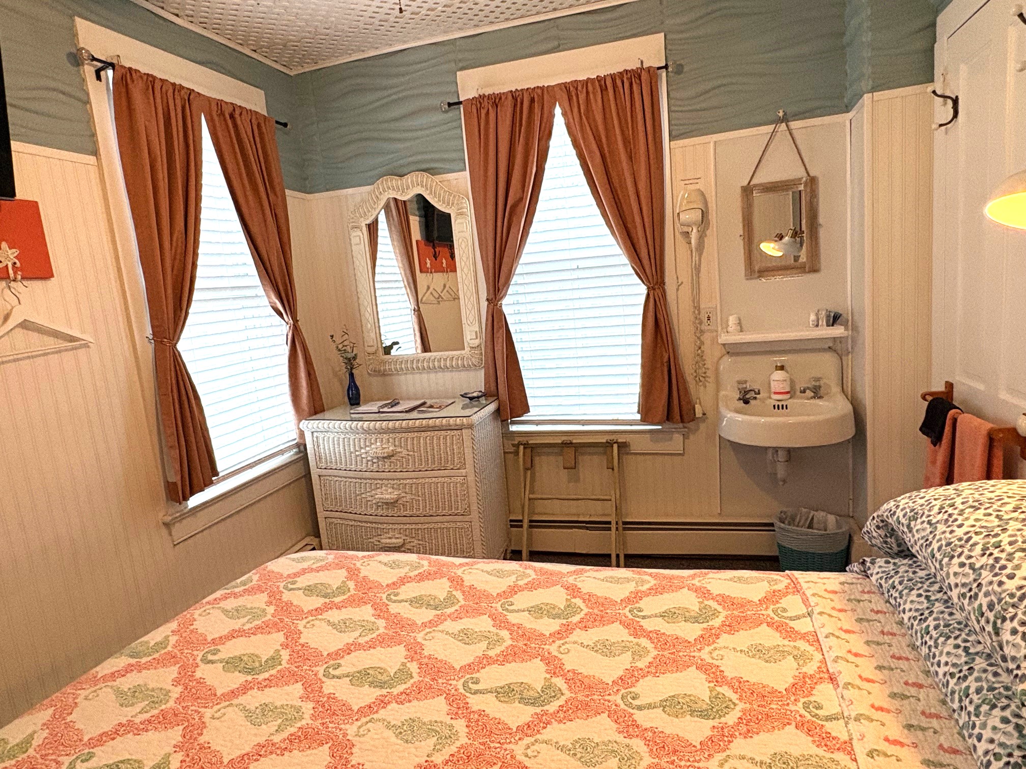 An interior view of a bedroom featuring a bed with a white wicker headboard and a coral-colored quilt, with two lit gooseneck lamps mounted on the blue wall above it.