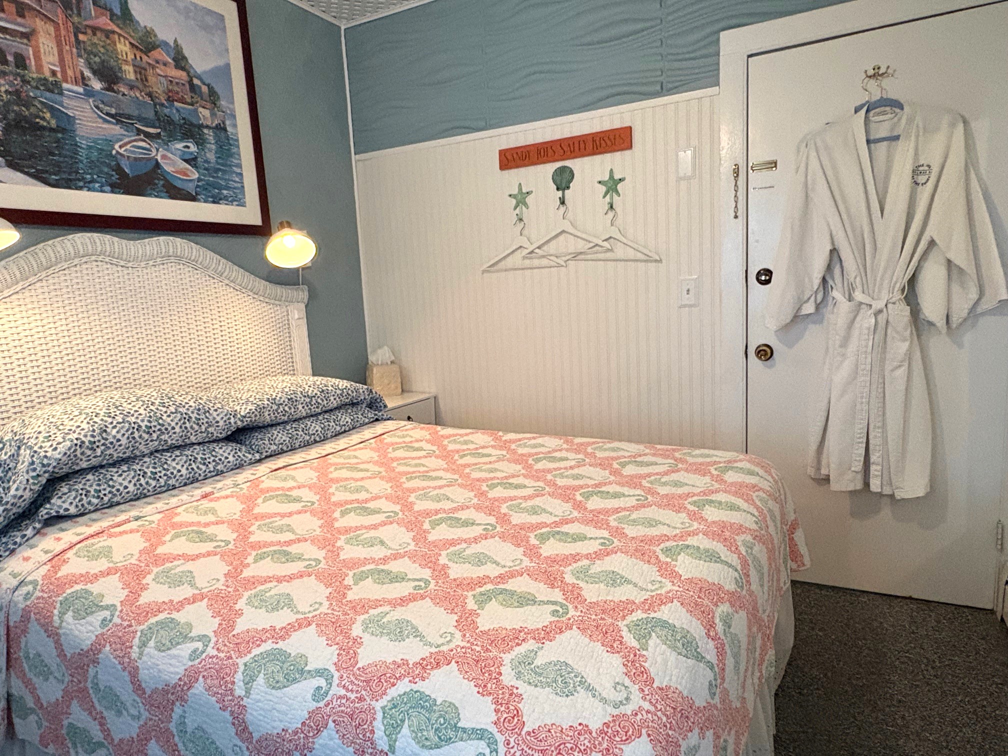 An interior view of a bedroom featuring a bed with a white wicker headboard and a coral-colored quilt, with two lit gooseneck lamps mounted on the blue wall above it.