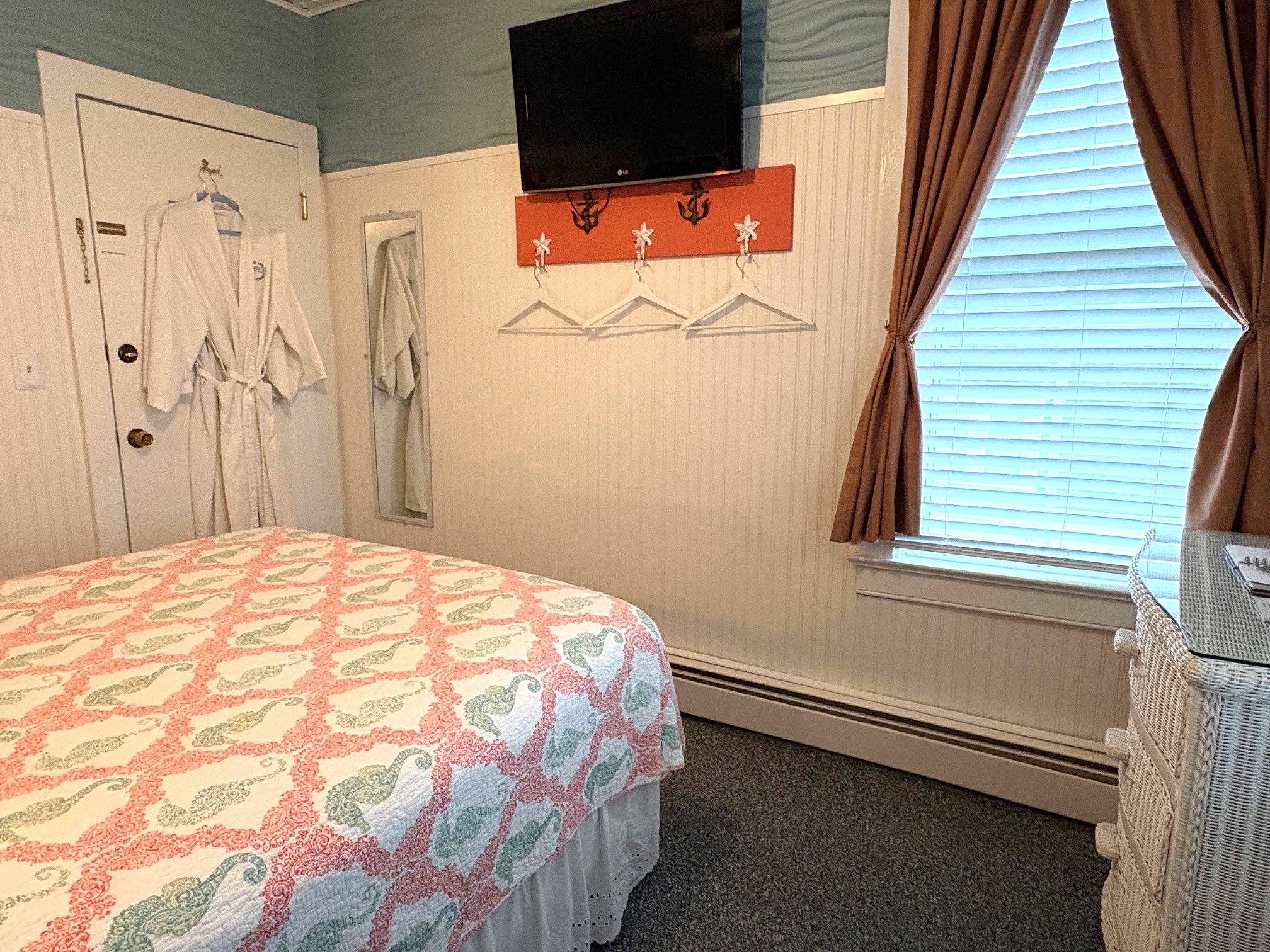 An interior view of a compact bathroom featuring a white pedestal sink, a white wicker-framed mirror, and a shower with a coral and seashell patterned curtain. A nautical anchor-shaped mirror hangs on the wall, and a stack of fresh white towels is visible to the left.