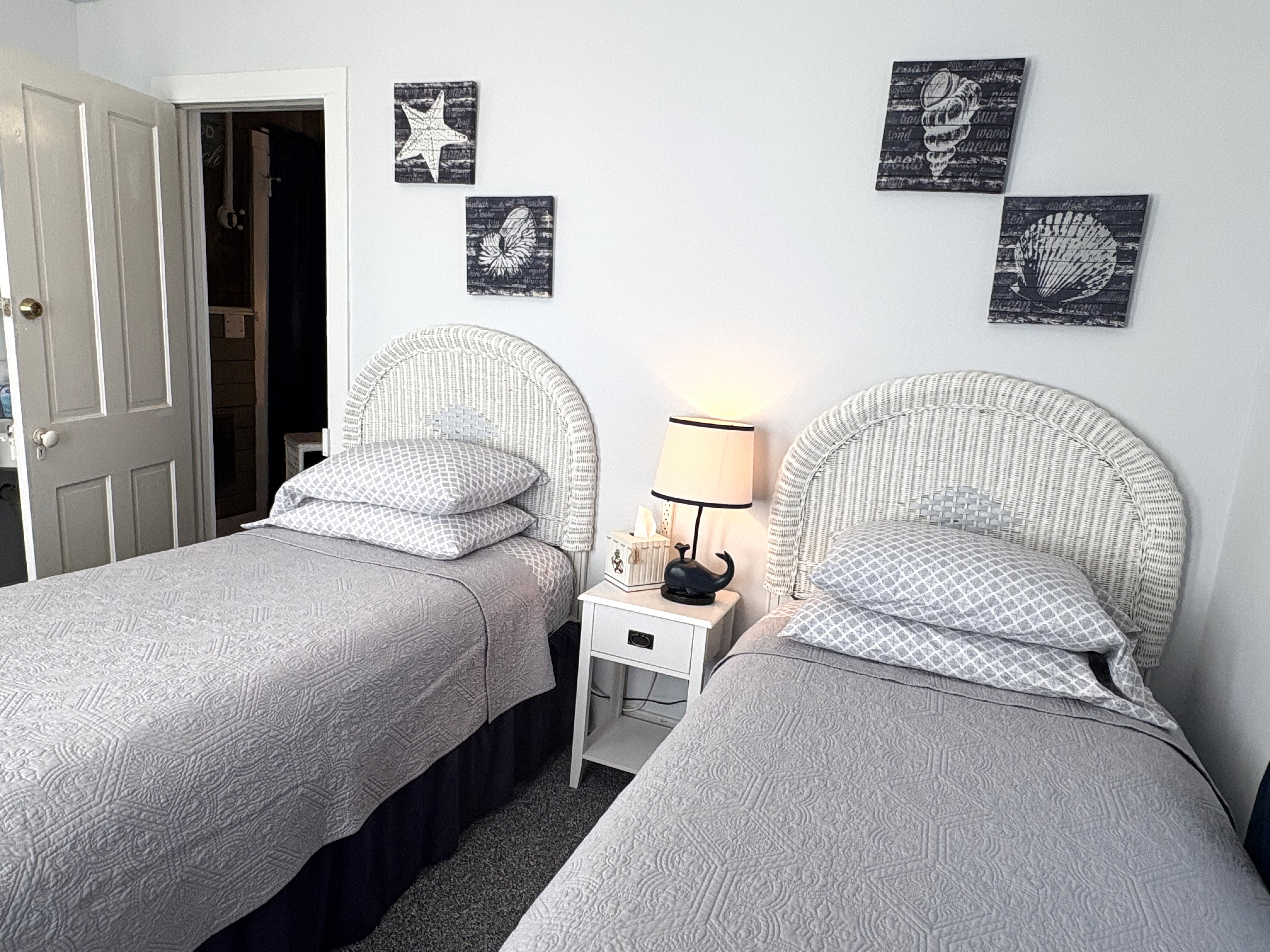 An interior view of a bedroom featuring two twin beds with white wicker headboards and gray quilted bedding. A white nightstand between the beds holds a whale-themed lamp, and nautical-themed starfish and seashell artwork hangs on the wall above the beds.