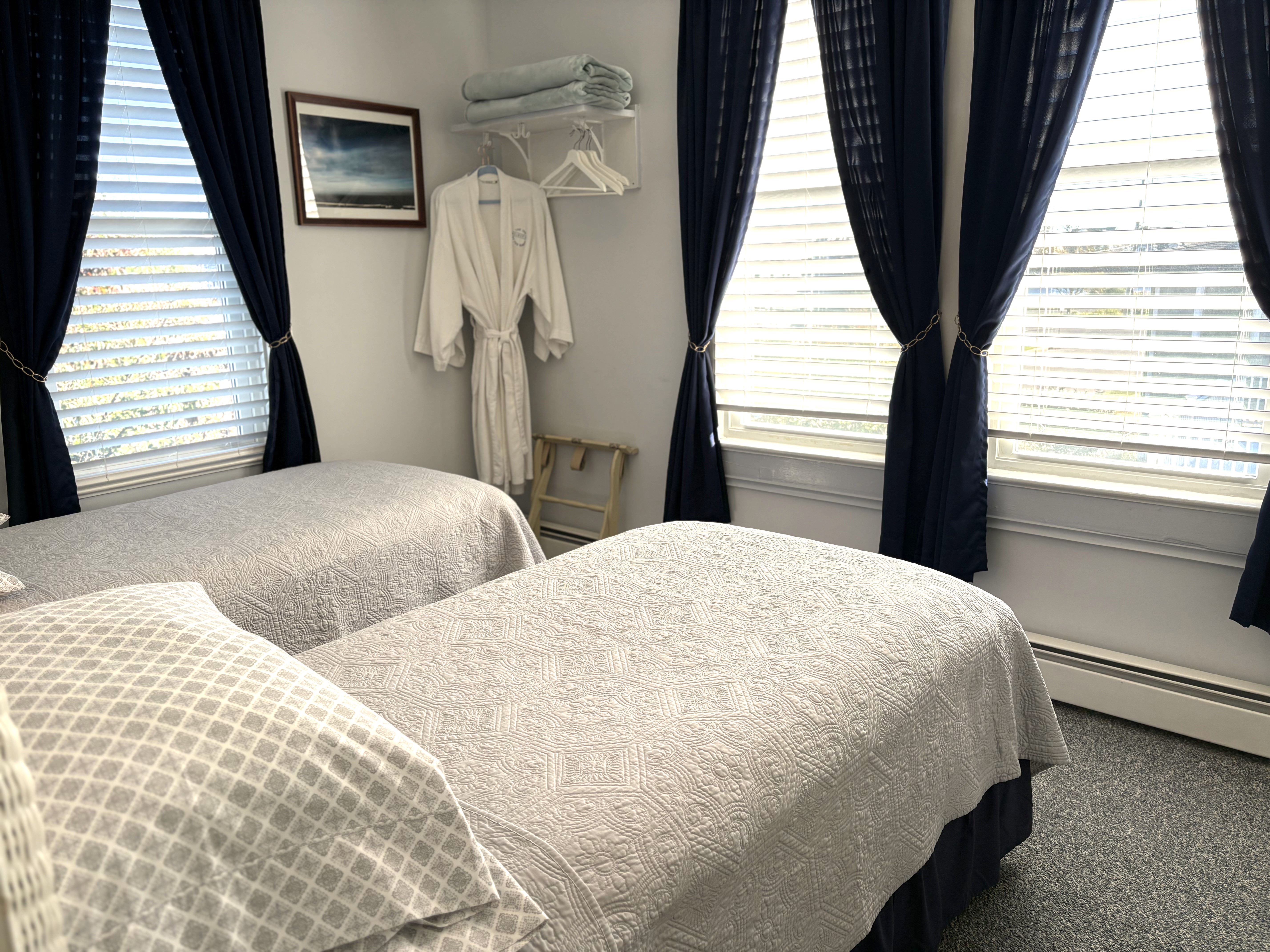 An interior view of a bedroom featuring two twin beds with white wicker headboards and gray quilted bedding. The room includes a white nightstand with a whale-themed lamp, navy blue curtains, and nautical wall art, including framed starfish and seashell prints.