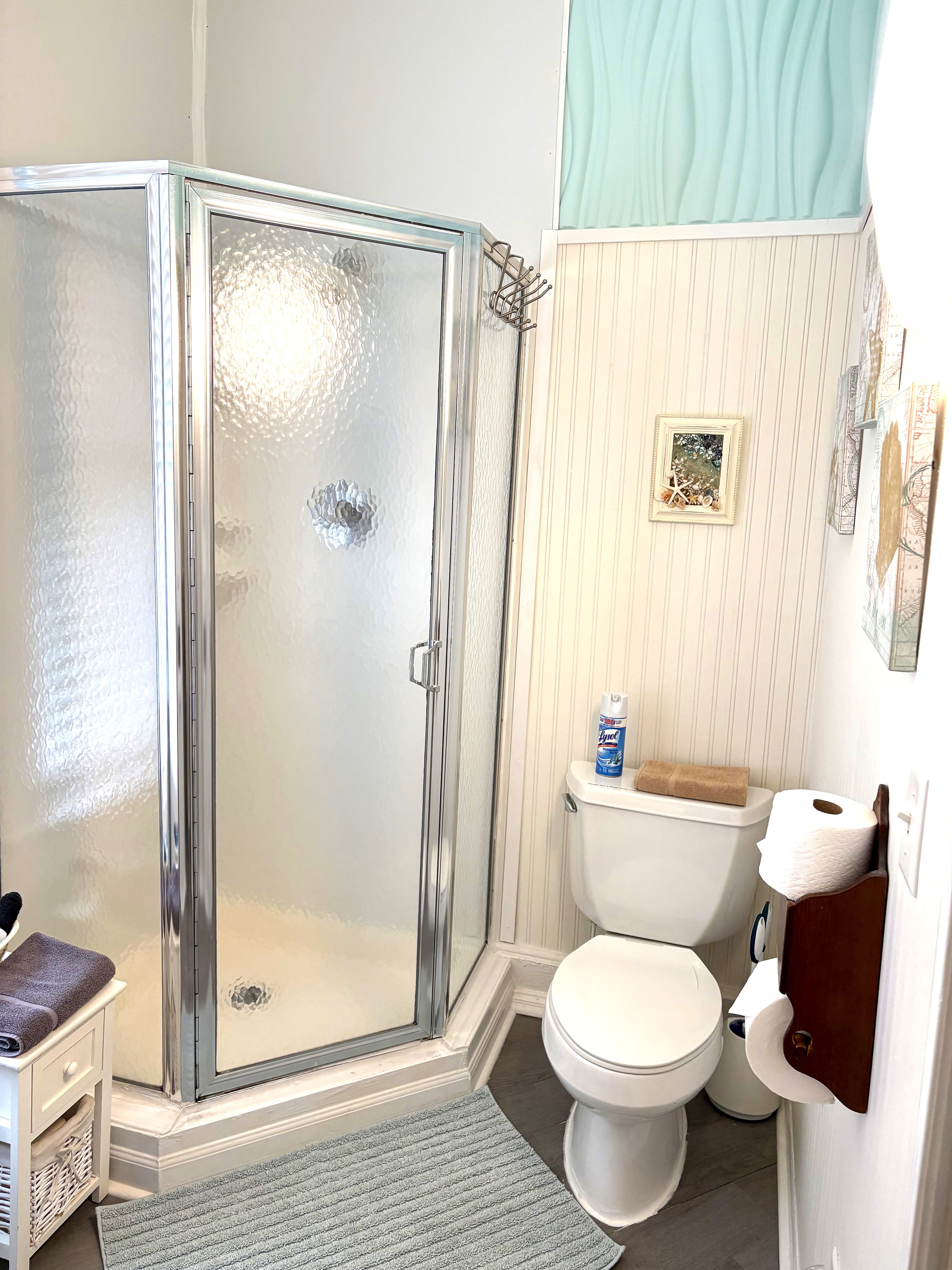 A bright en-suite bathroom featuring a corner walk-in shower with a glass door, a white toilet, and white beadboard walls decorated with small framed coastal art.