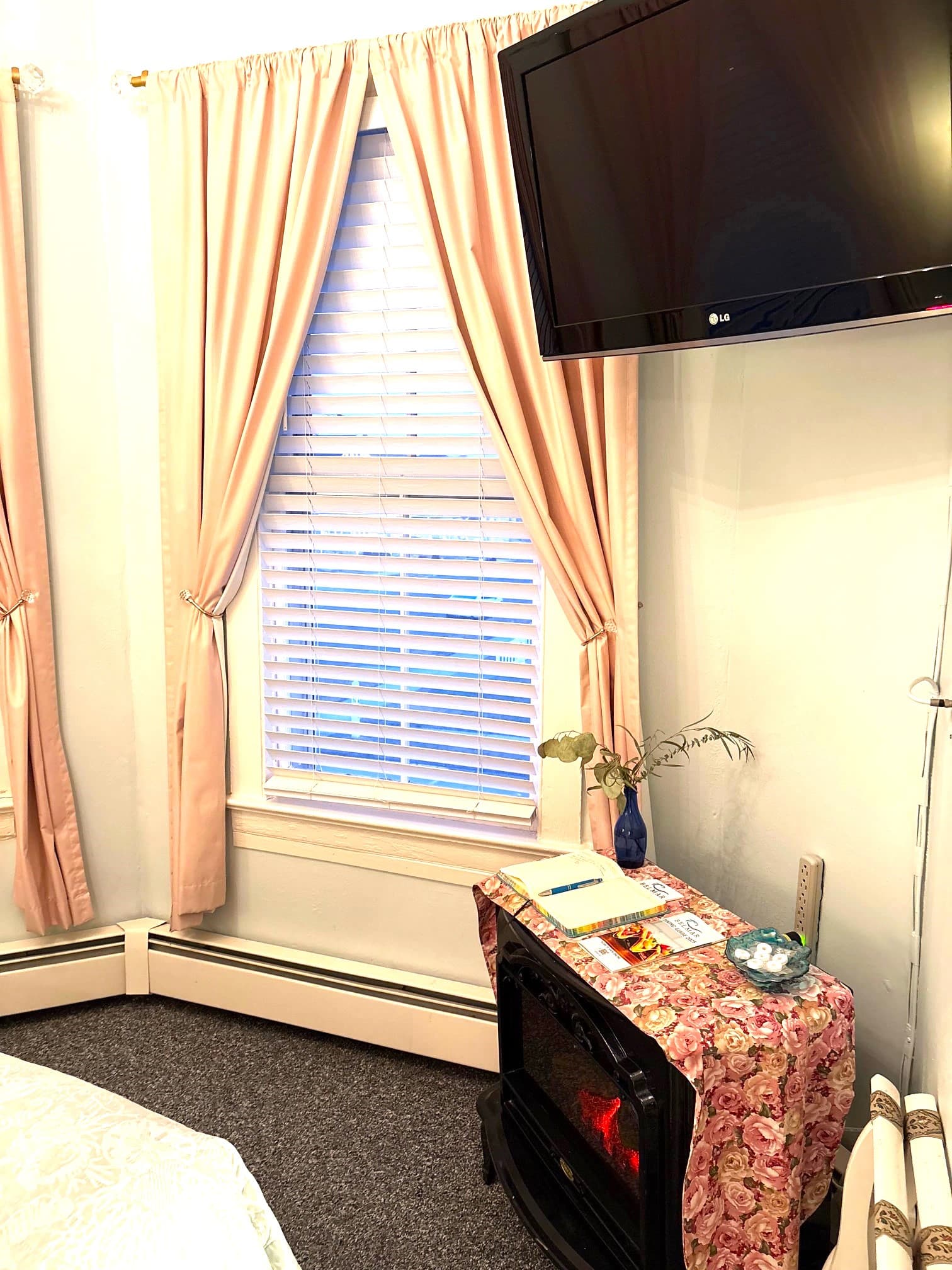 An interior view of a bathroom featuring a classic white claw-foot tub with a shower curtain, a toilet, and an open door with a full-length mirror. A small area with patterned wallpaper and a shelf is visible to the right.