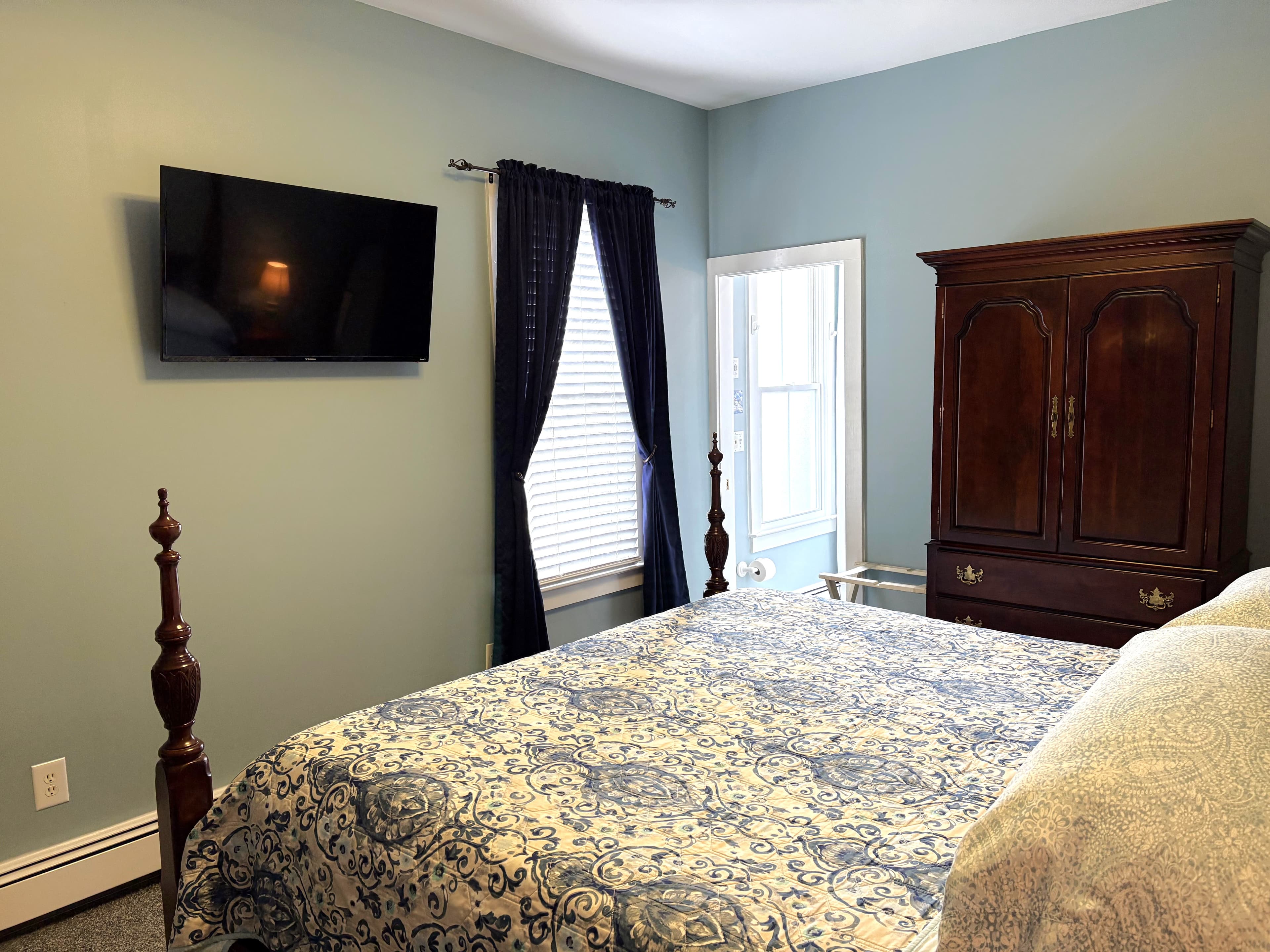 An interior view of a bedroom featuring a dark wood four-poster bed with patterned blue and white bedding, a large dark wood armoire, and a wall-mounted television.