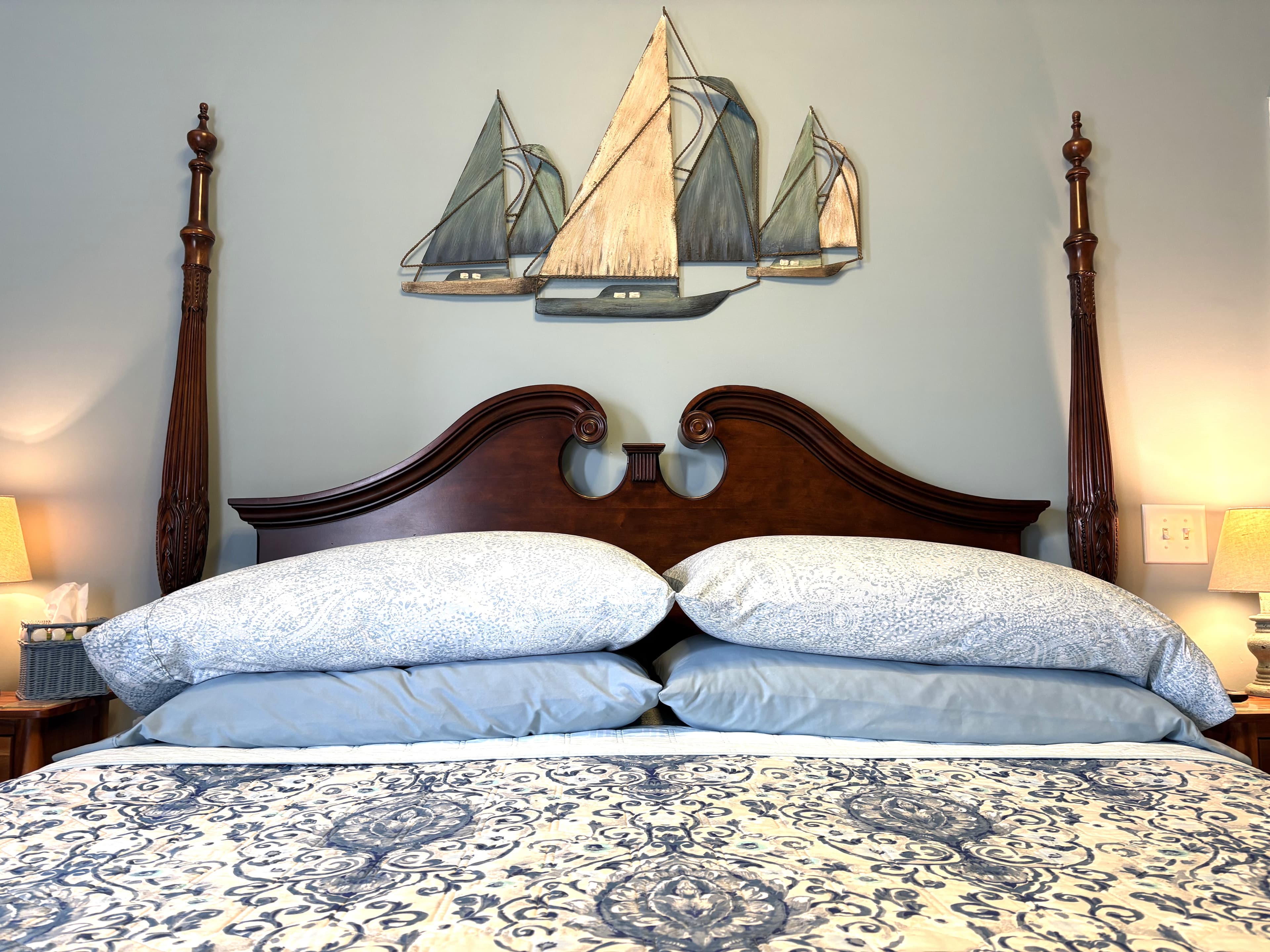 An interior view of a bedroom featuring a dark wood four-poster bed with a curved headboard, a blue and white patterned comforter, and a large metal wall hanging of three sailboats above the bed.