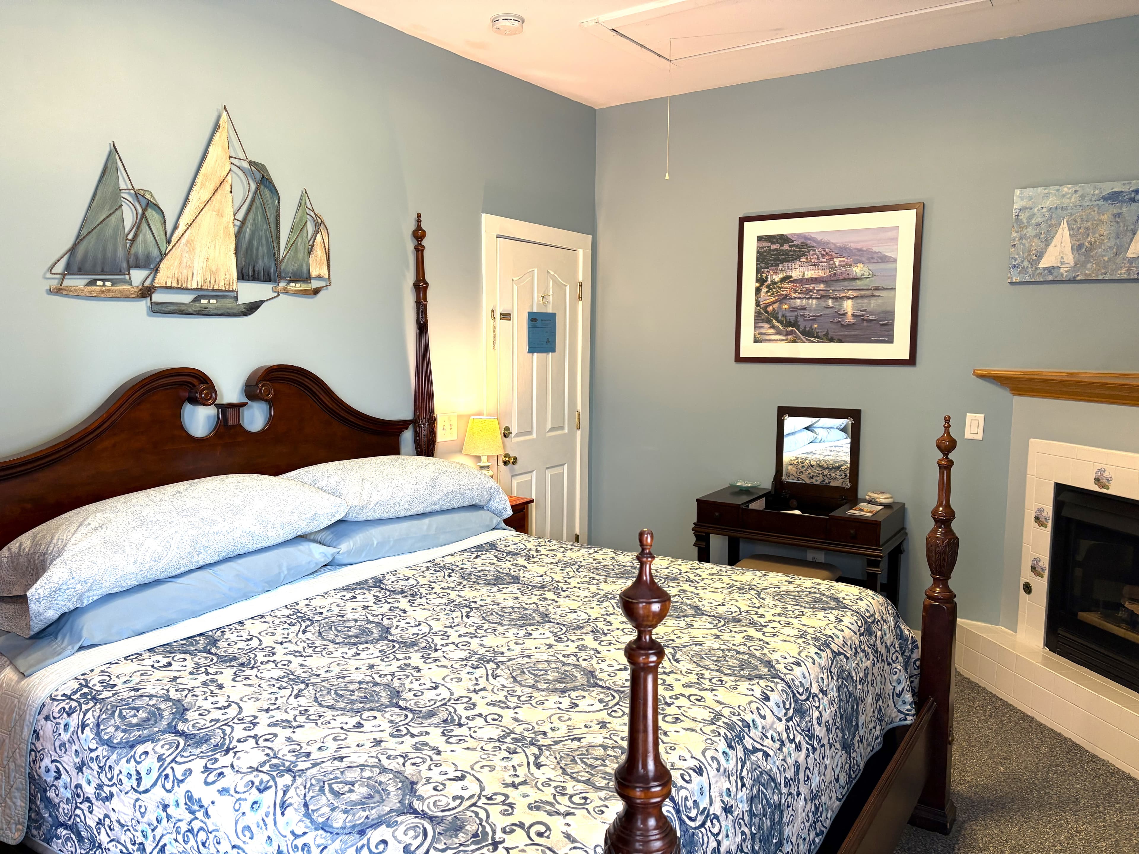 An interior view of a spacious bedroom featuring a dark wood four-poster bed with blue and white patterned bedding. To the right, a built-in gas fireplace is visible, along with a desk and several pieces of coastal-themed wall art, including a large sailboat sculpture above the bed.
