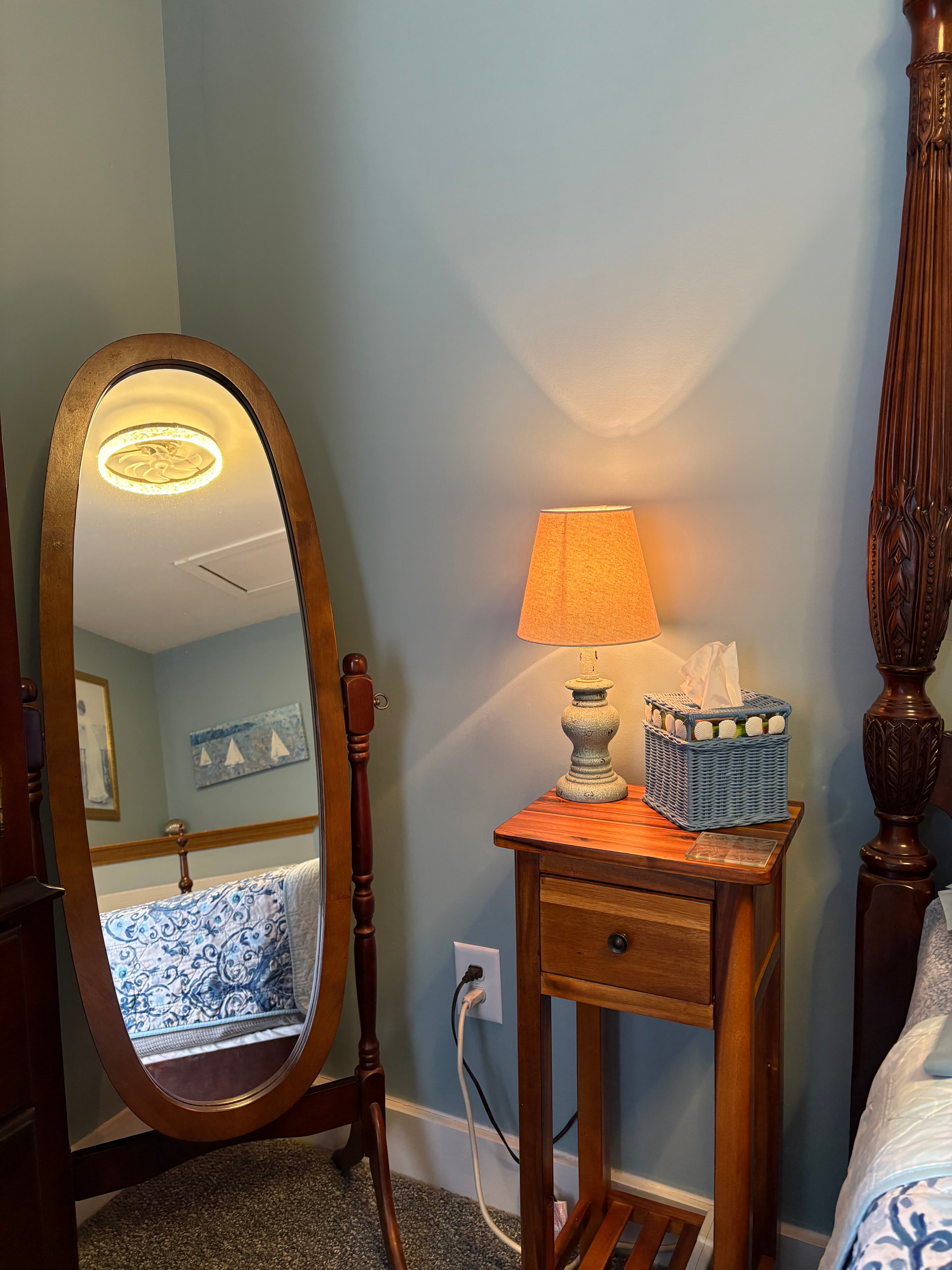 An interior view of a bedroom corner featuring a full-length oval dressing mirror on a wooden stand, an antique-style wooden nightstand with a lit lamp, and the post of a dark wood bed frame.