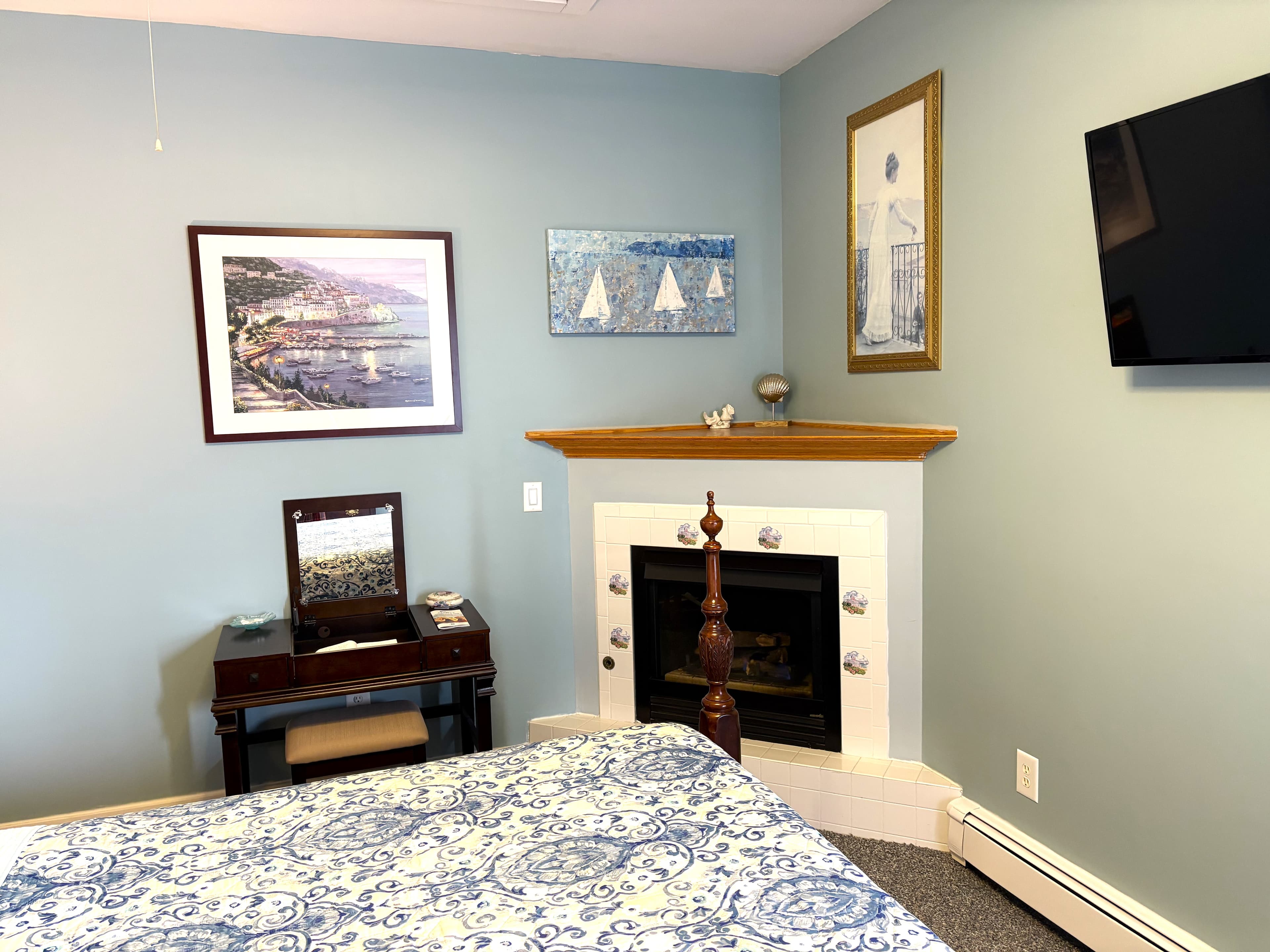 An interior view of a bedroom corner featuring a built-in corner fireplace with a white mantel and a small desk with a vanity mirror and stool. Several coastal-themed framed pictures are displayed on the light blue walls.