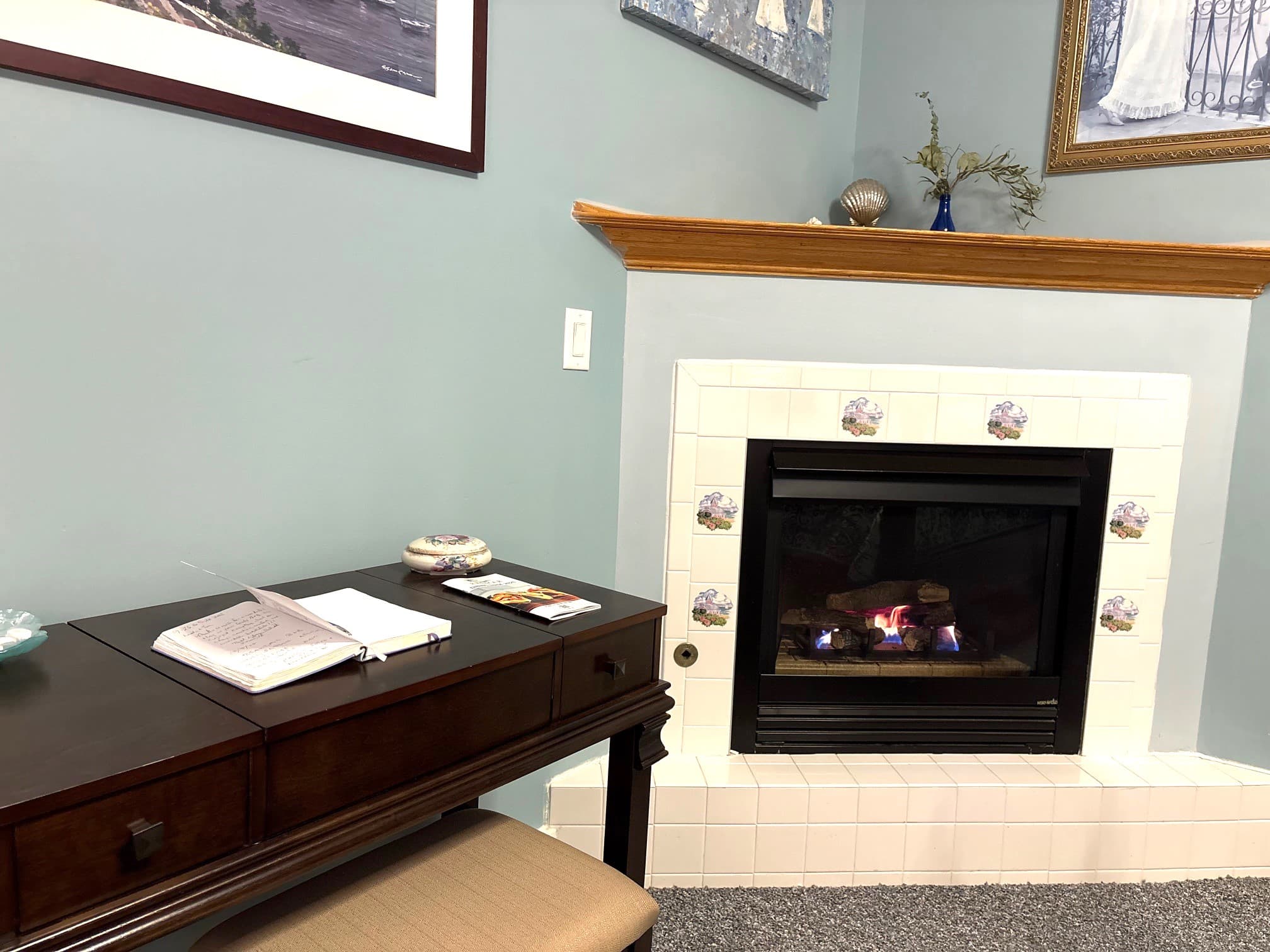 A close-up of a lit built-in gas fireplace featuring realistic logs and a warm glow, surrounded by a white tile hearth accented with decorative tiles.