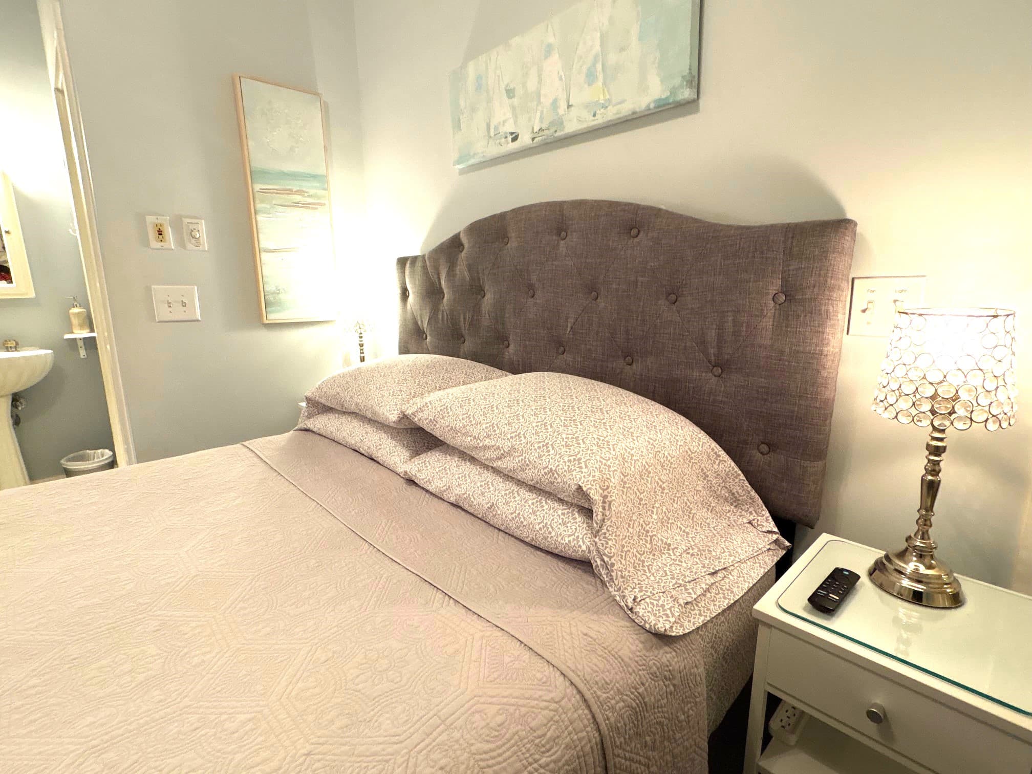 An interior view of a bedroom featuring a plush, grey tufted headboard with white and grey patterned bedding. Two decorative crystal lamps are lit on white bedside tables, and a horizontal coastal-themed painting hangs on the wall above the bed.