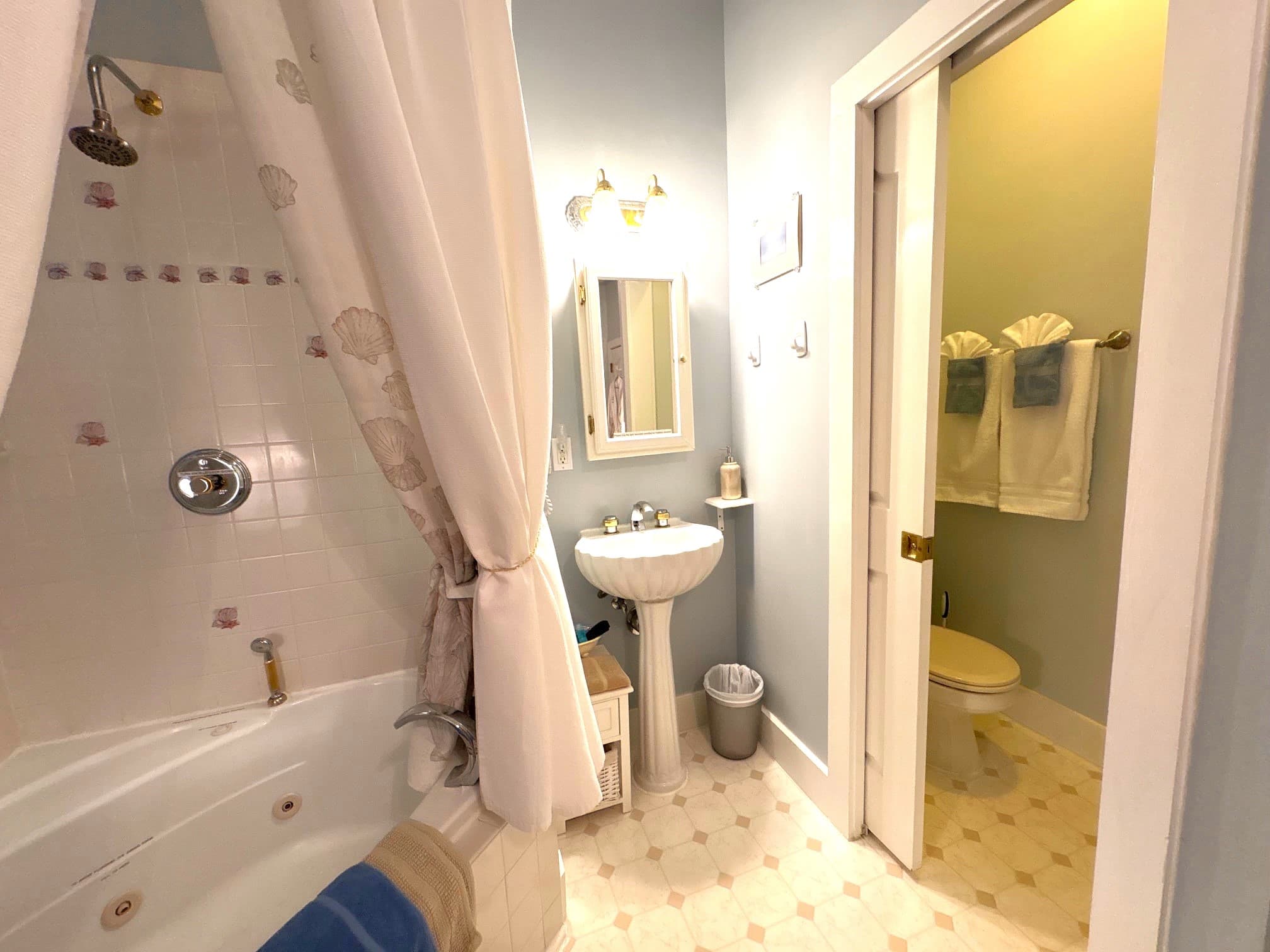 An interior view of a bright guest room featuring a plush gray tufted headboard and a bed with a circular patterned quilt. To the right, a built-in corner fireplace with a white mantel is visible, and an open doorway leads to a bathroom with a white pedestal sink and a lit mirrored cabinet.