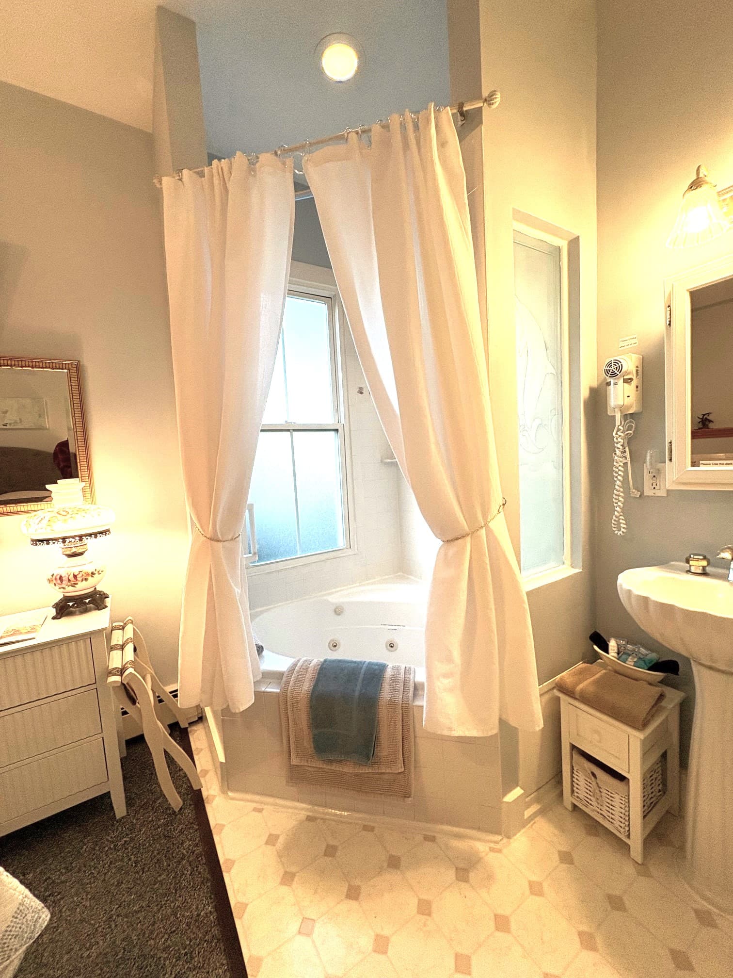 An interior view of a bright guest room featuring a plush gray tufted headboard, a bed with a circular patterned quilt, a ceiling fan, and a built-in corner fireplace with a white mantel and decorative tiles.