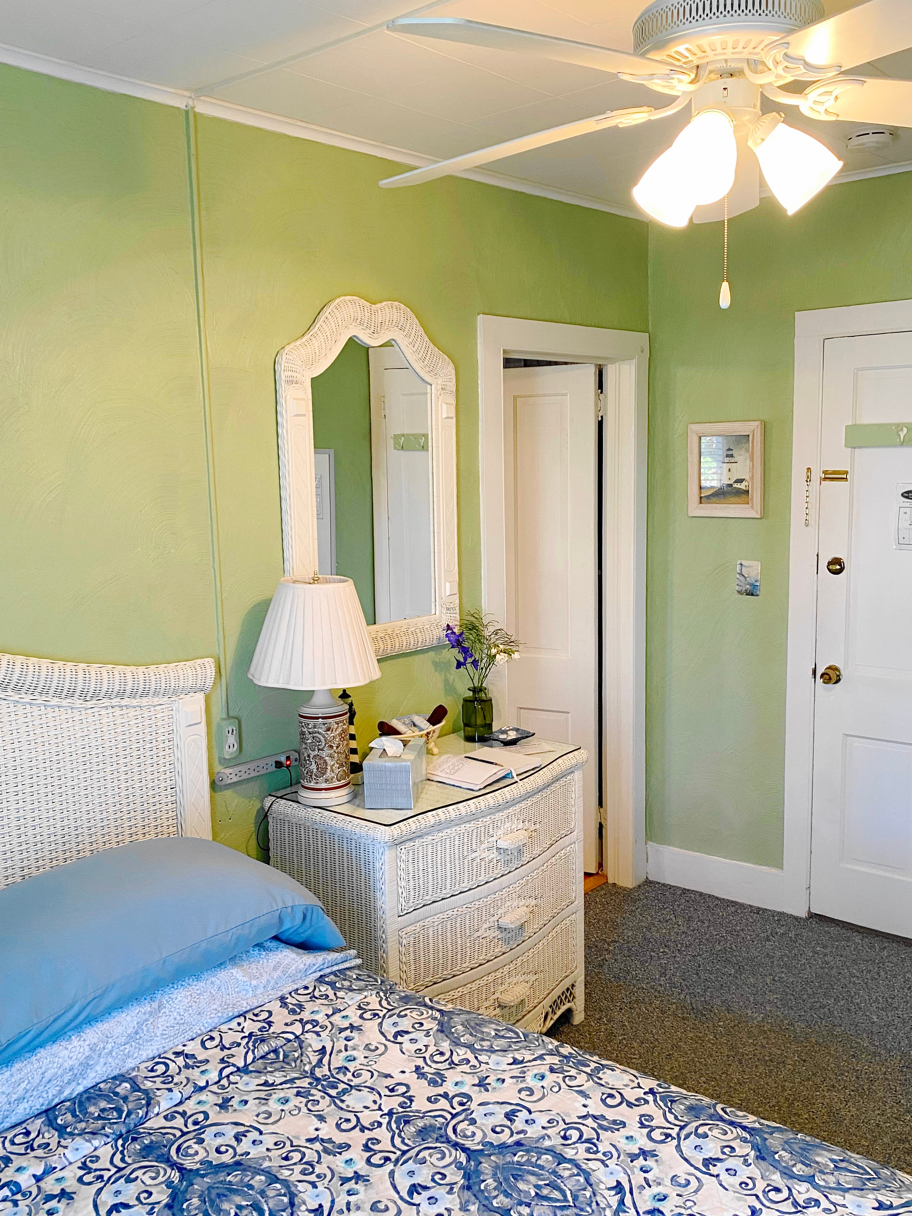 An interior view of a bedroom with light green walls featuring a bed with a white wicker headboard and blue patterned bedding. A small white wicker wall shelf hangs above the bed next to a piece of coastal wall art depicting flip-flops on a sandy beach.