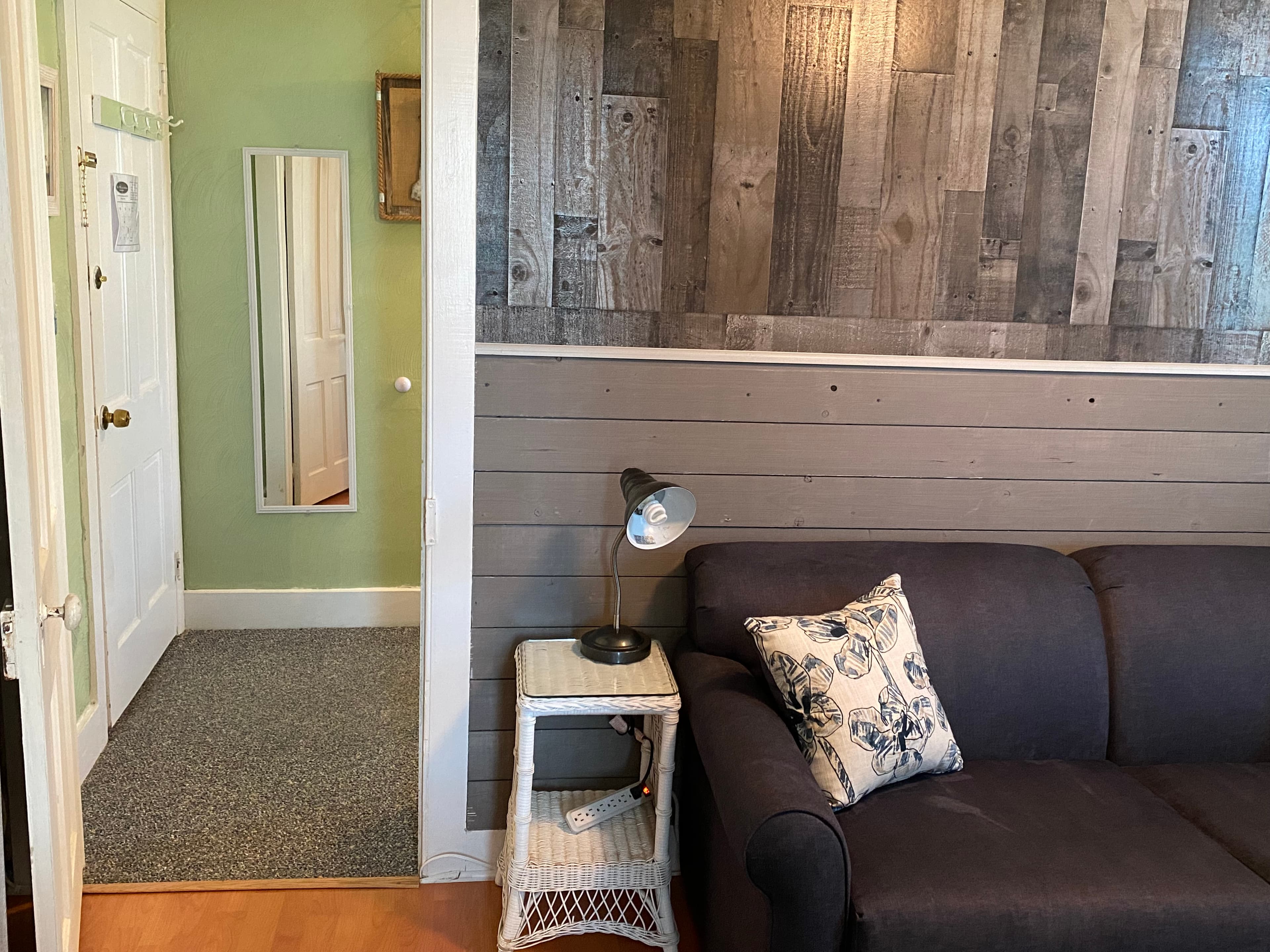 An interior view of a cozy seating area featuring a dark gray sofa with decorative floral pillows, a traditional blue and gray patterned rug, and a small bistro-style table with two stools. A wall-mounted television is positioned across from the sofa, and a lit ceiling fan provides a warm glow over the wood-plank flooring.