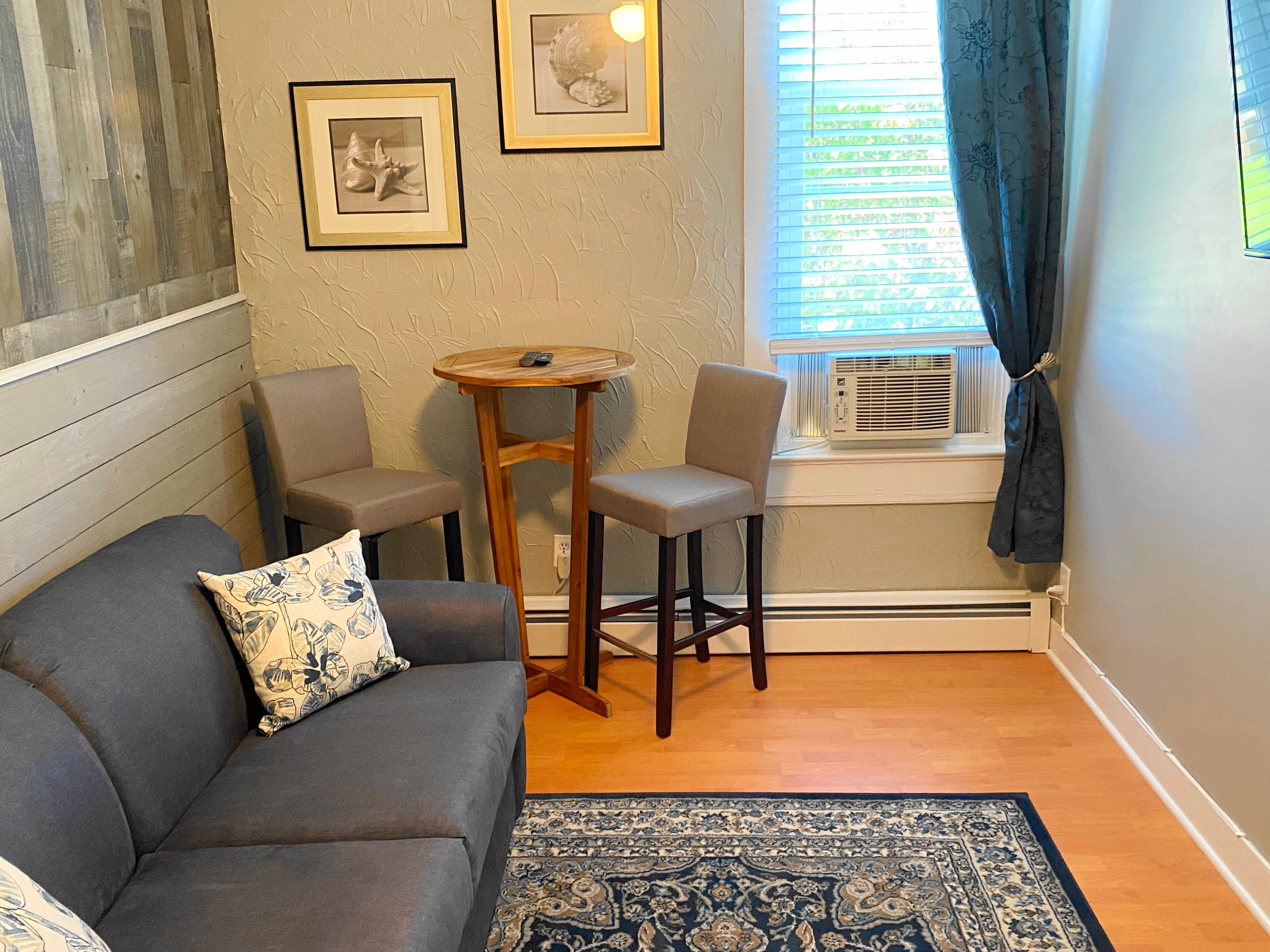 An interior view of a cozy seating area featuring a black leather wingback armchair, a gray louvered folding privacy screen, and a small white wicker side table set against light-colored walls and wood-plank flooring.