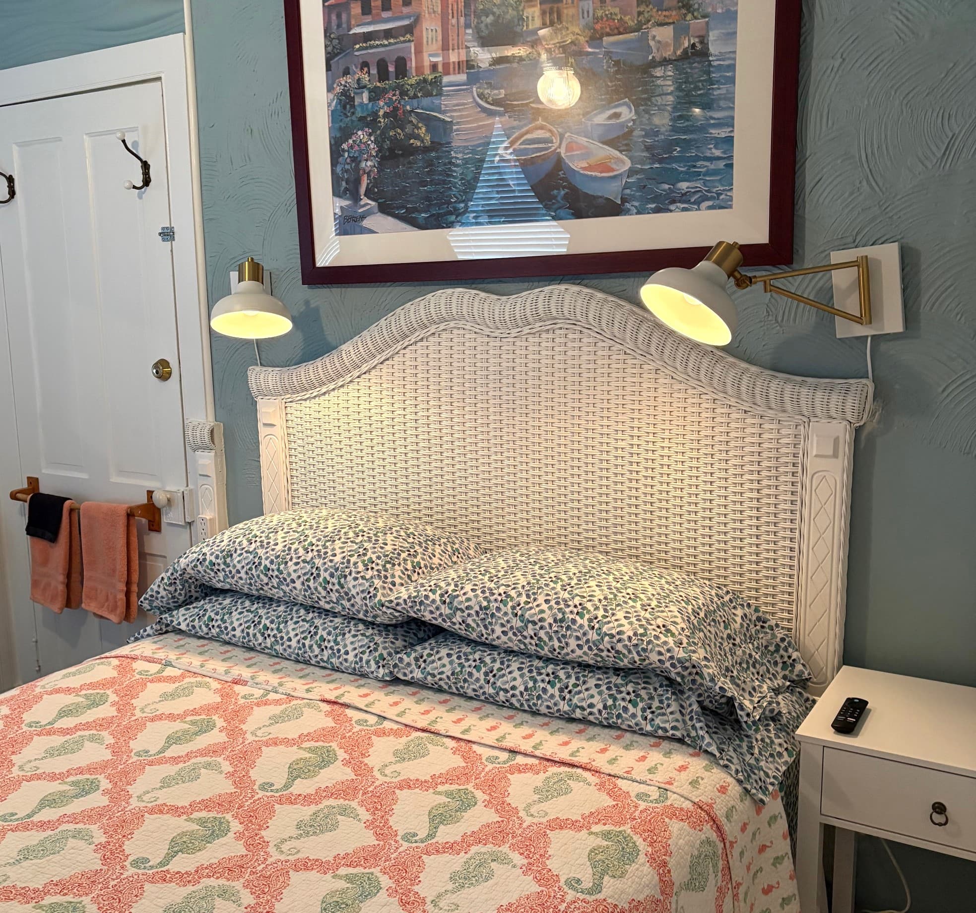 An interior view of a bedroom featuring a white beadboard headboard with two lit, wall-mounted reading lamps and a bed covered in a coral-colored textured quilt.