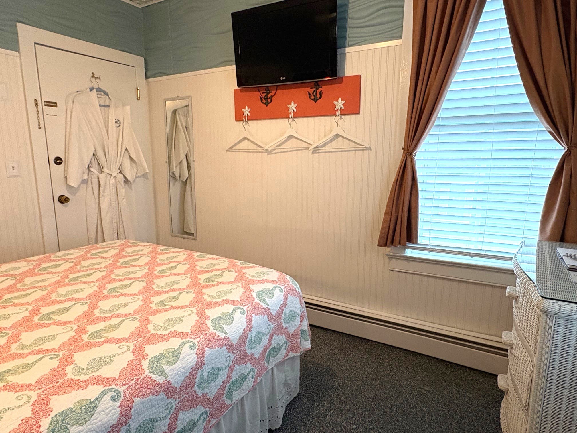 An interior view of a compact bathroom featuring a white pedestal sink, a white wicker-framed mirror, and a shower with a coral and seashell patterned curtain. A nautical anchor-shaped mirror hangs on the wall, and a stack of fresh white towels is visible to the left.