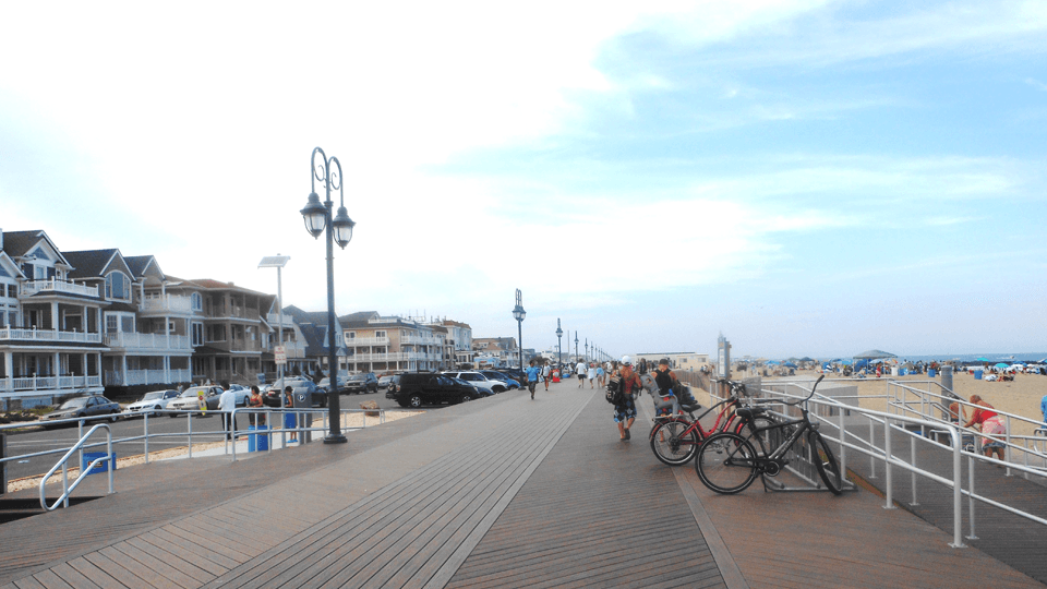 A wooden boardwalk lined with houses, bicycles, and pedestrians leading to the beach under a cloudy sky.