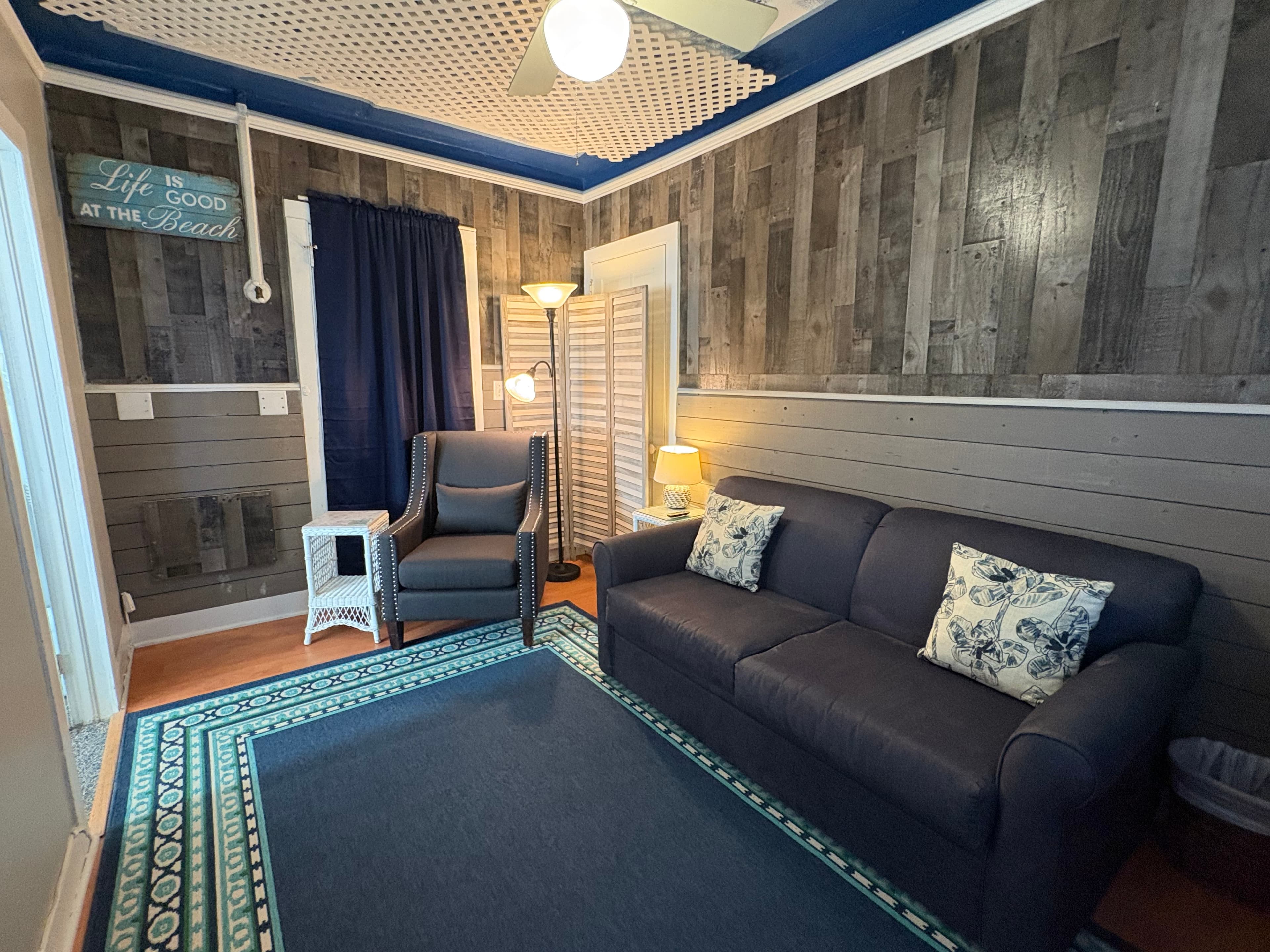 Cozy living room with a dark wood-paneled wall, a navy blue sofa, and a chair, featuring warm lighting and beach-themed decor.