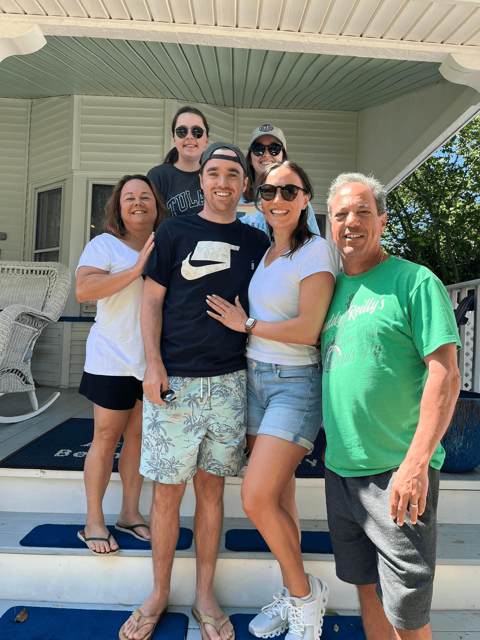 A group of six people pose together on a porch, smiling and enjoying a sunny day.