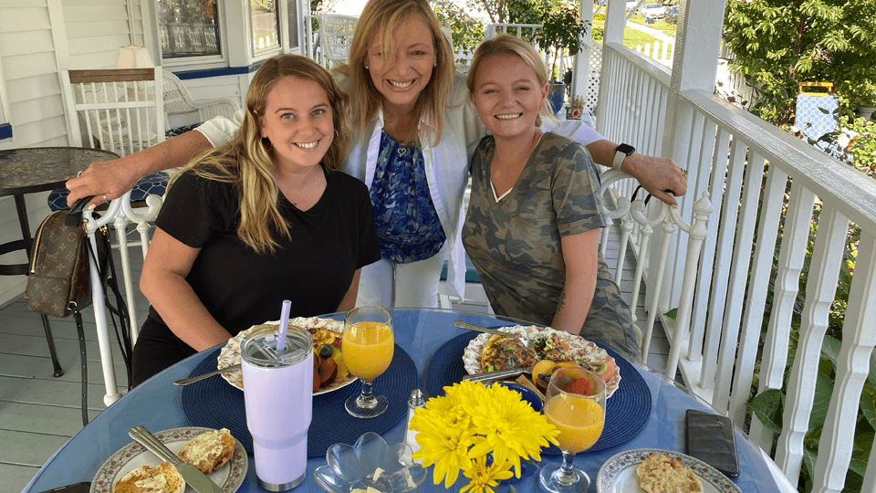 Three women enjoy a meal together on a porch, surrounded by flowers and dishes.