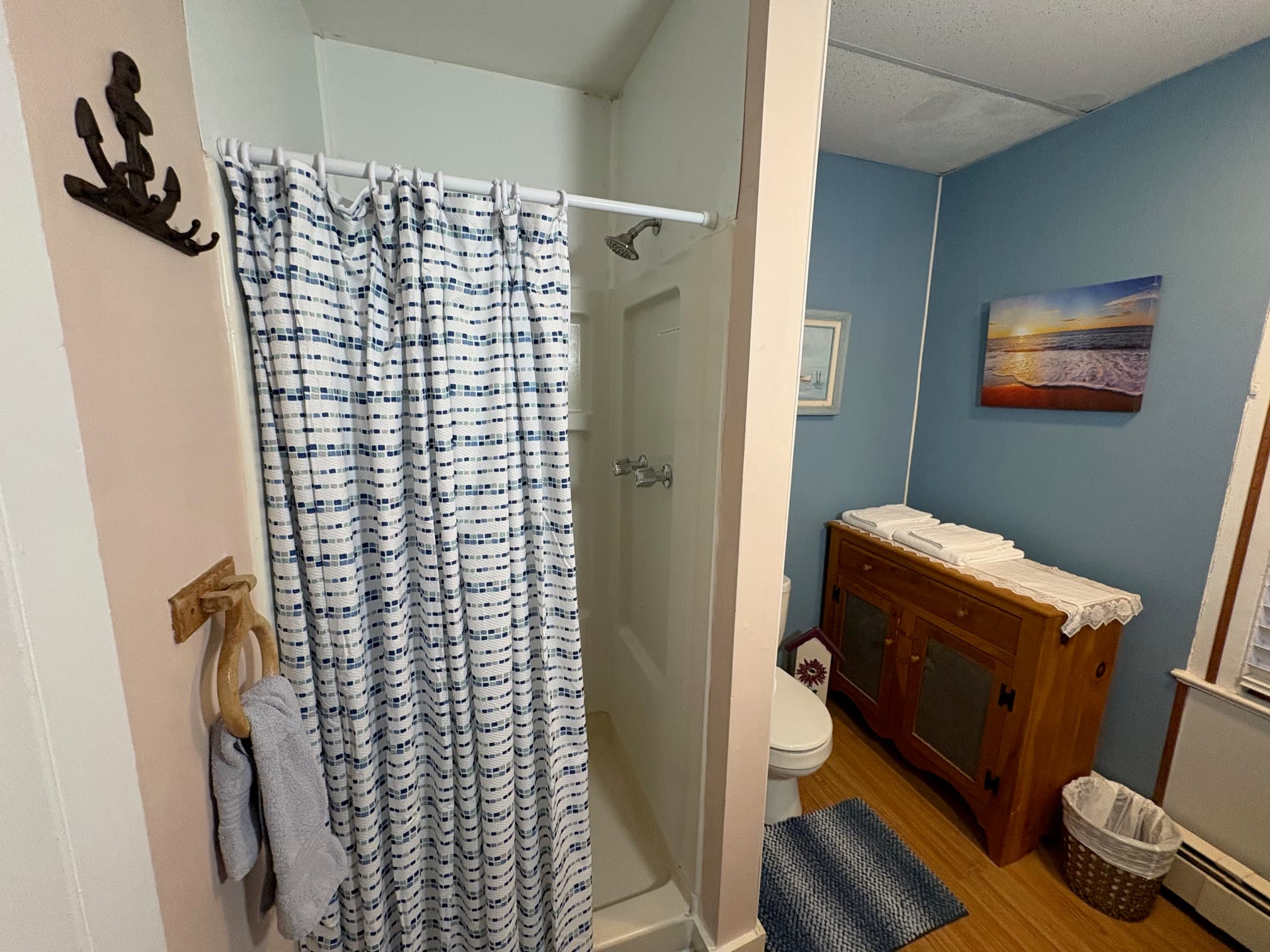 A small bathroom featuring a shower with a blue and white curtain, a toilet, a wooden cabinet, and coastal-themed artwork on the walls.