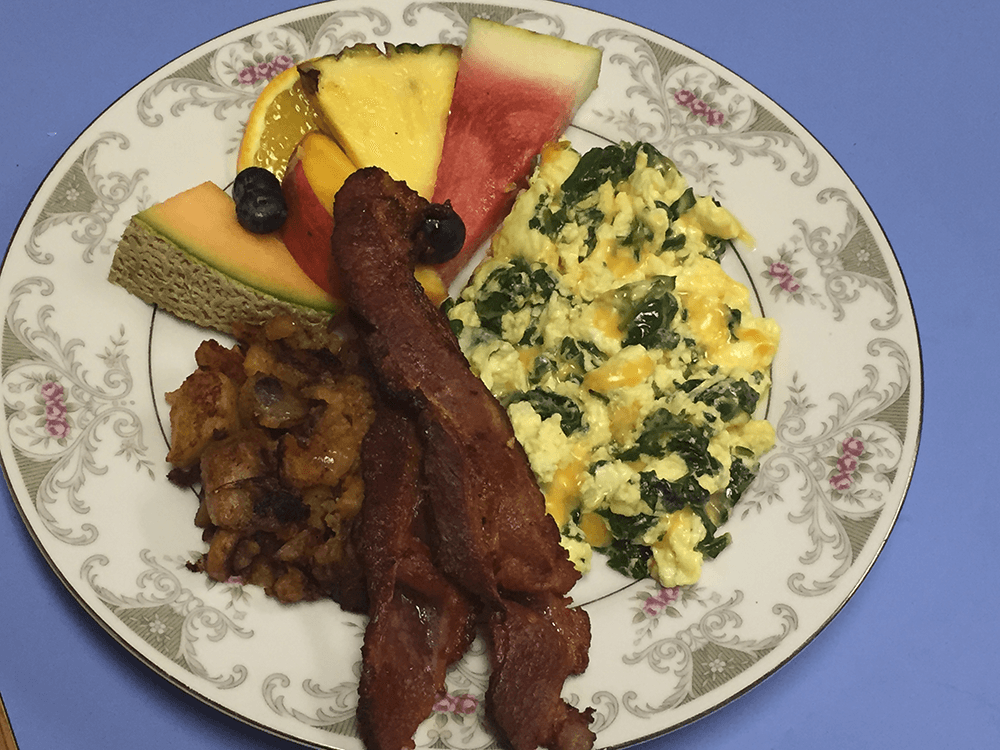 A plate of scrambled eggs with spinach, crispy bacon, breakfast potatoes, and assorted fruit slices.