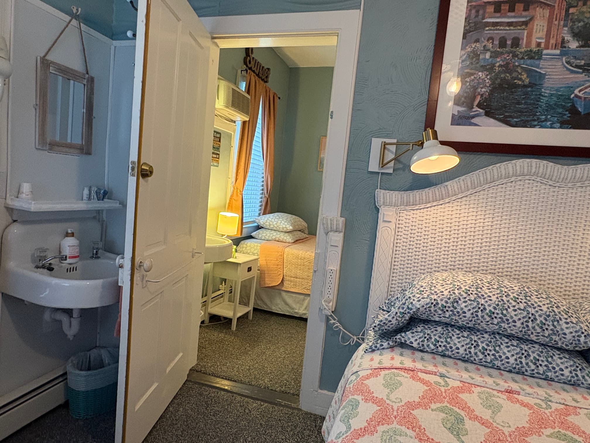 A cozy bedroom with a white wicker headboard, featuring a small sink, and a doorway leading to an adjacent room with two beds.