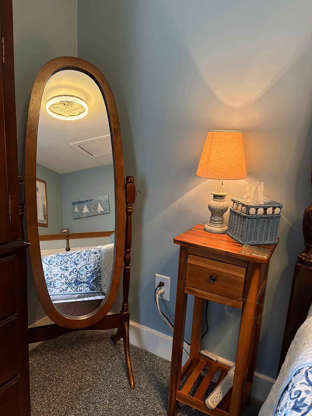 A wooden framed mirror stands next to a bedside table with a lamp and a tissue box, in a softly lit bedroom with blue walls.