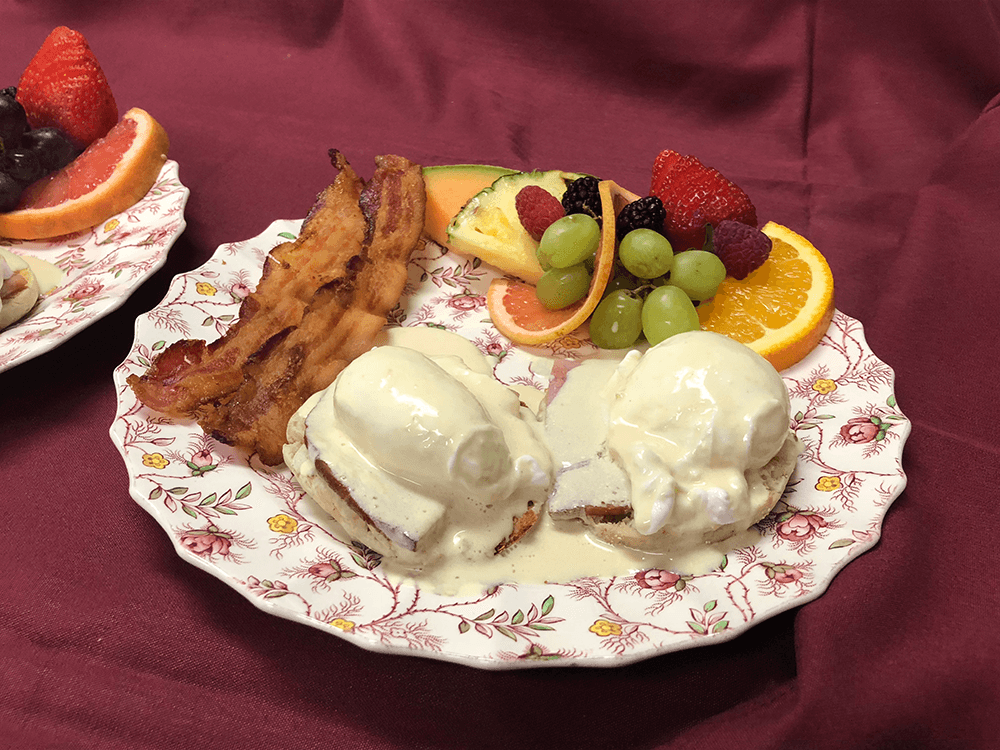 A plate of eggs Benedict topped with hollandaise sauce, accompanied by bacon and a colorful fruit medley.