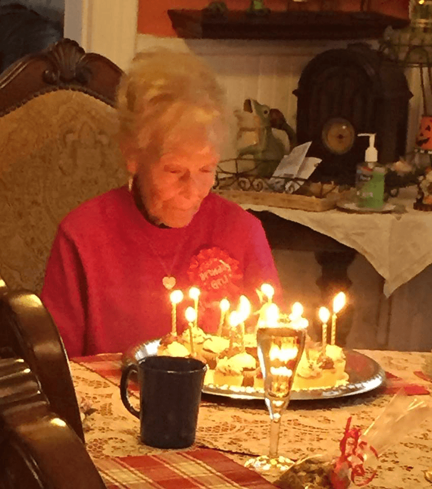 An elderly woman sits at a table with a plate of cupcakes adorned with lit candles, reflecting on the moment.