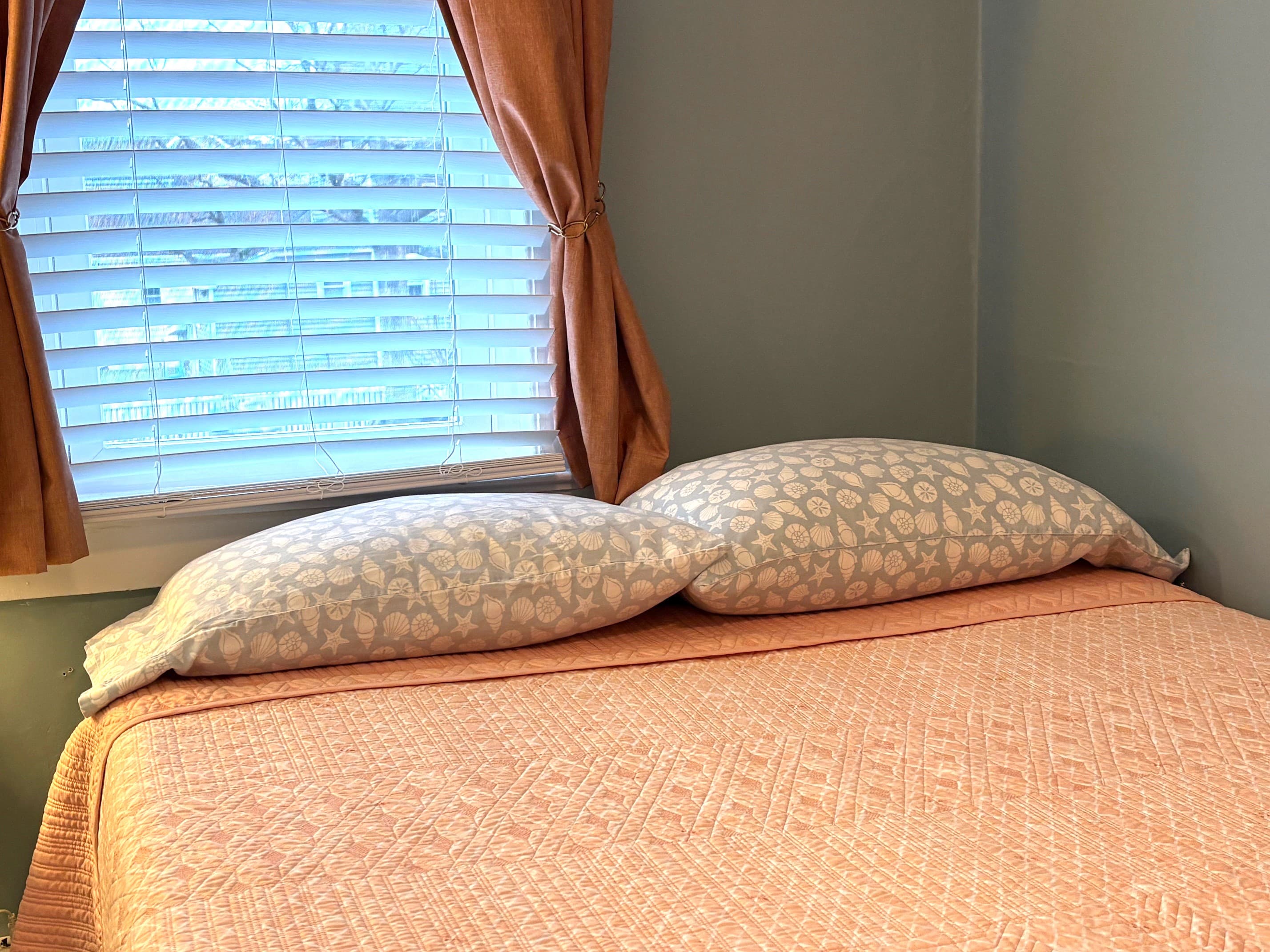 A neatly made bed with floral pillows and an orange quilt, beside a window with blinds.