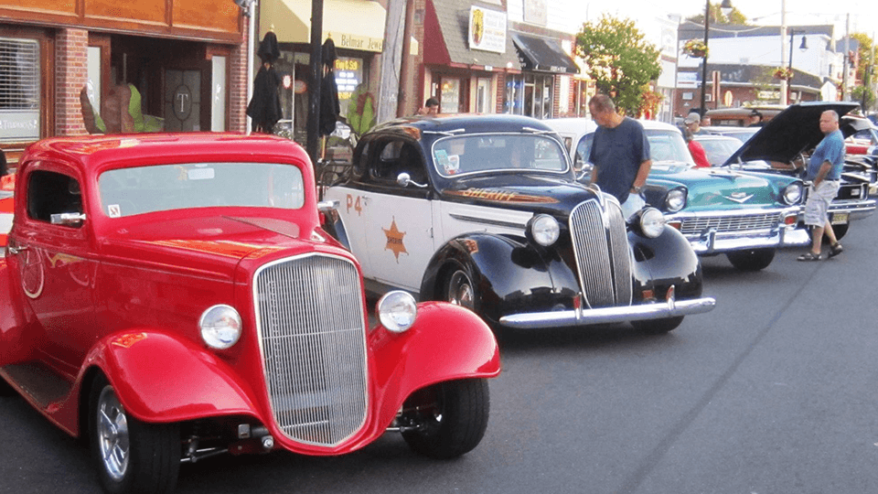 A row of classic cars parked on a street, with people inspecting them.