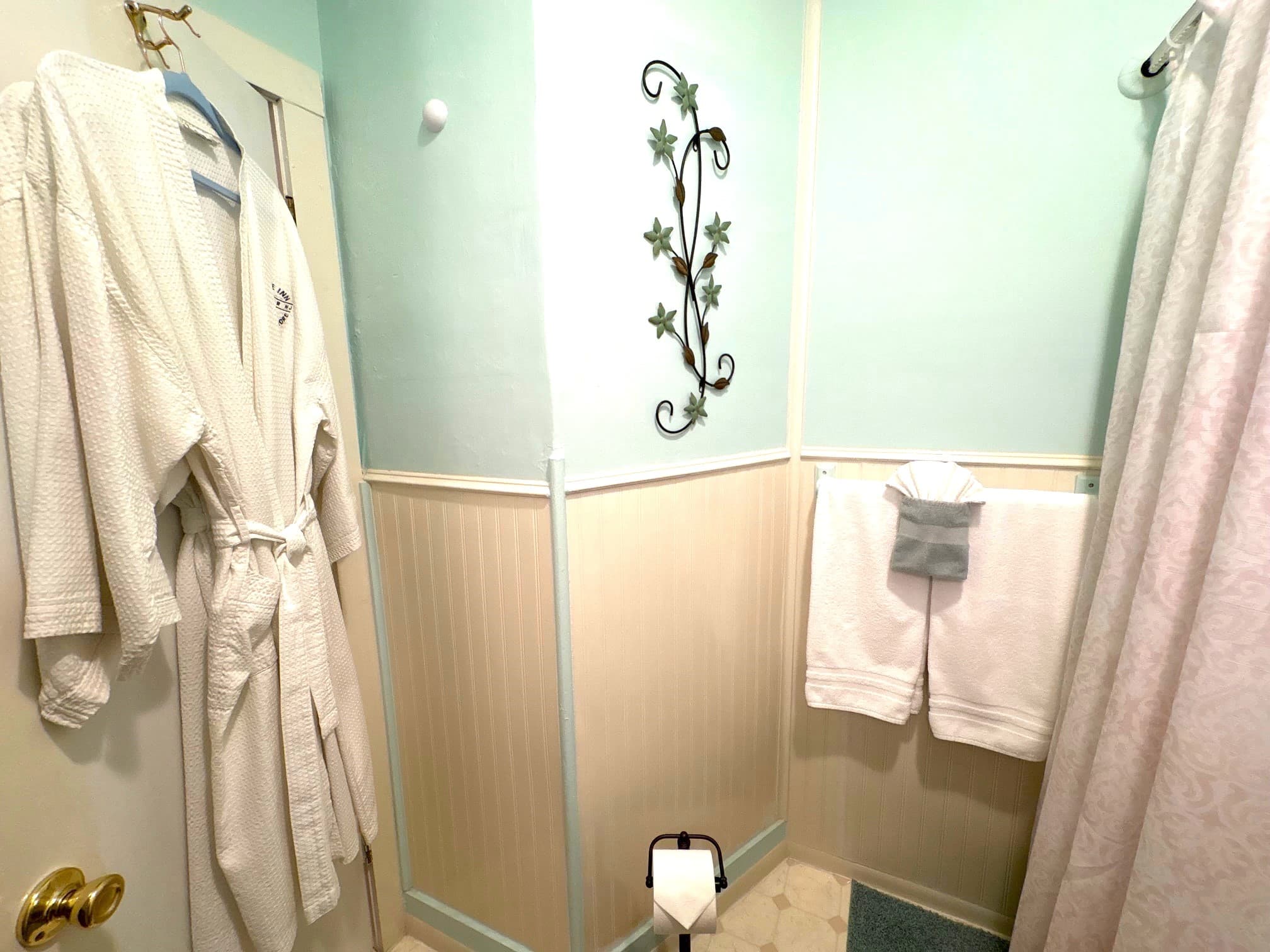 A cozy bathroom featuring light teal walls, a decorative wall vine, bathrobes, and neatly hung towels.