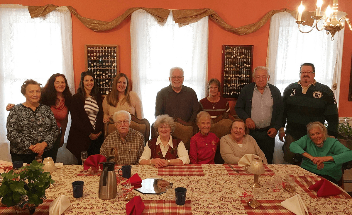 A diverse group of twelve people poses together around a dining table set for a meal in a warmly decorated room.