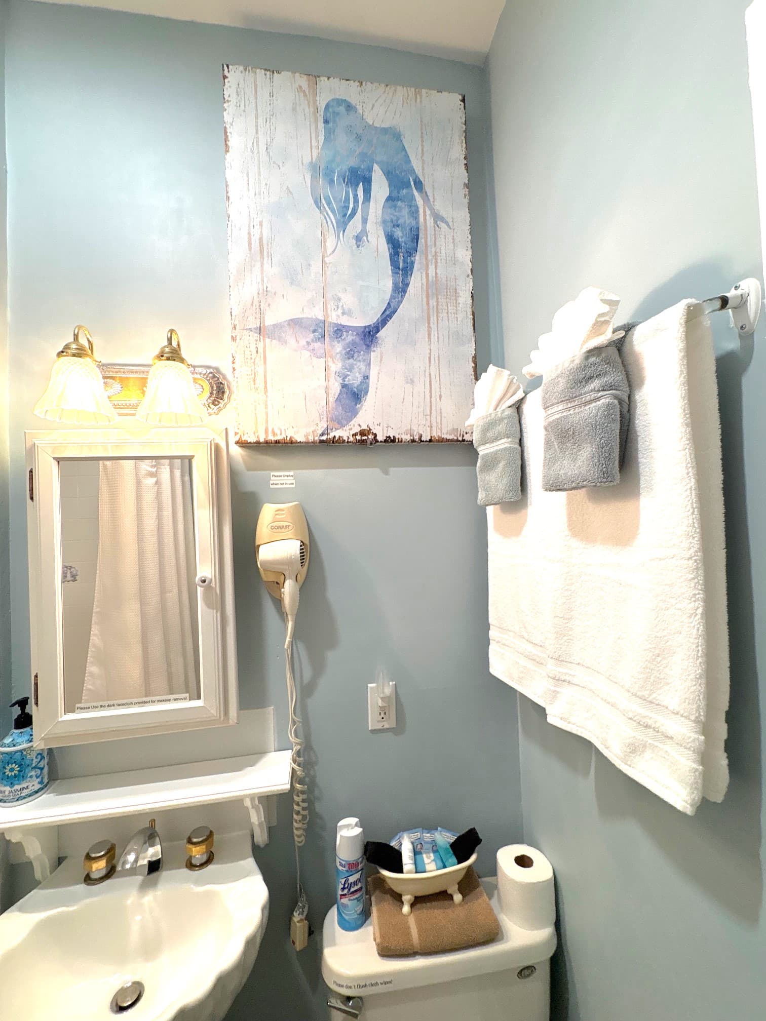 A small bathroom featuring a blue mermaid painting, a mirror, sink, and neatly arranged towels.